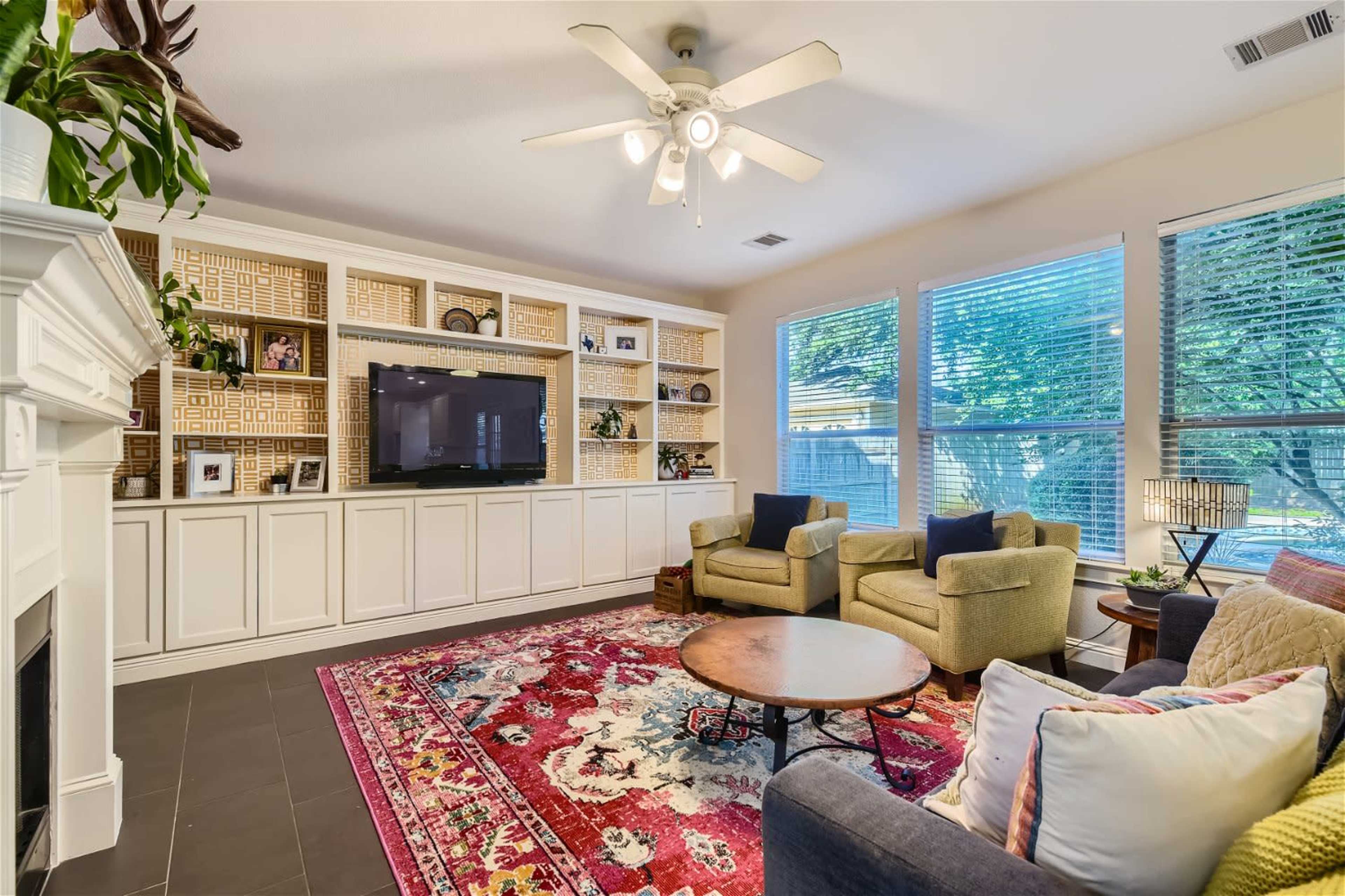A living room with a large flat-screen TV mounted above a built-in shelf, two armchairs, and a coffee table on a colorful area rug.