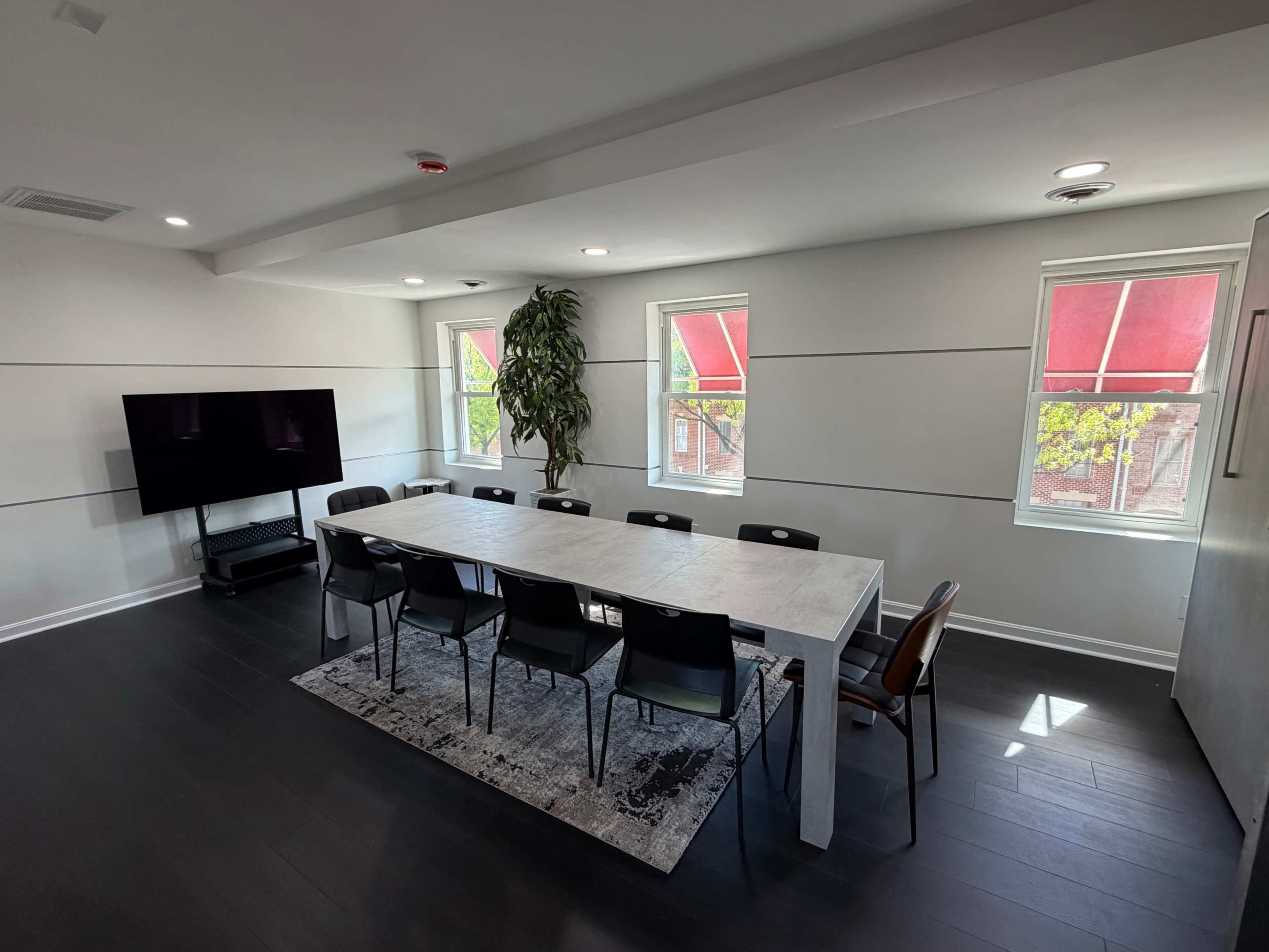 The image shows a modern conference room with a long white table surrounded by black chairs, large windows with red shades, and a TV mounted on the wall.