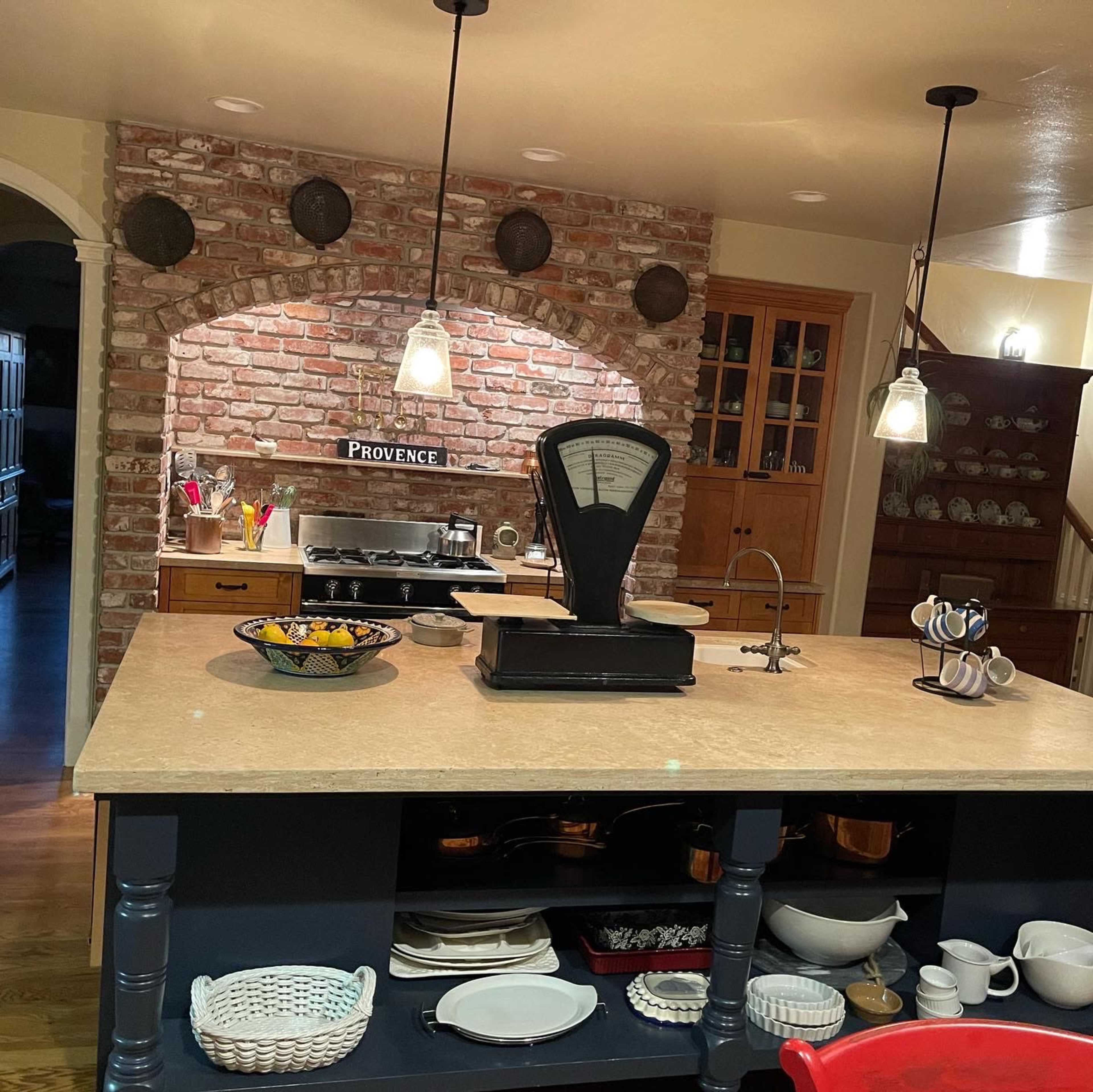 The kitchen features a central island with a scale, a brick wall with decorative plates, and wooden cabinetry.