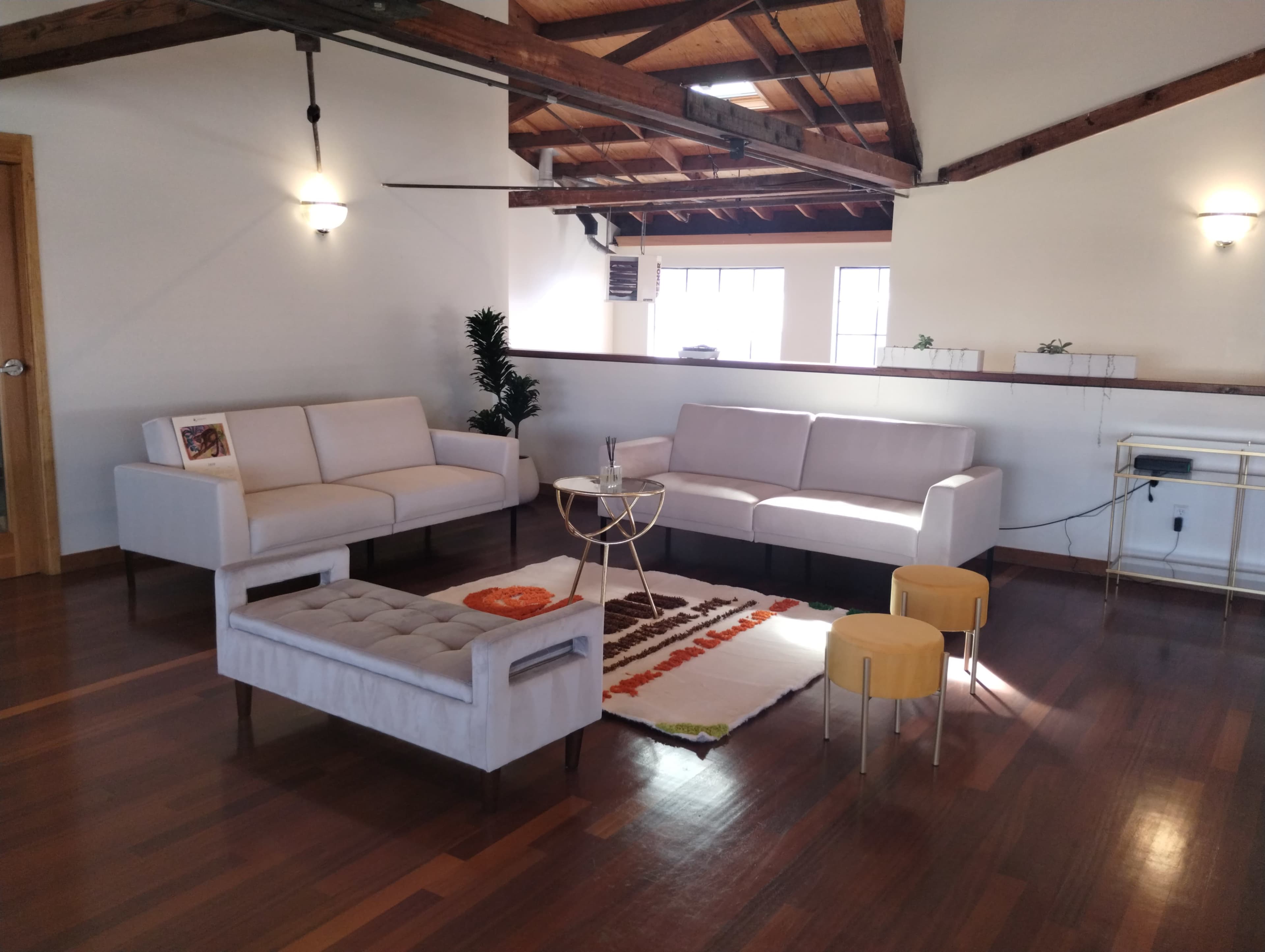 A spacious living area with two sofas, a coffee table, a patterned rug, and a few small stools, all set against a backdrop of wooden beams and large windows.