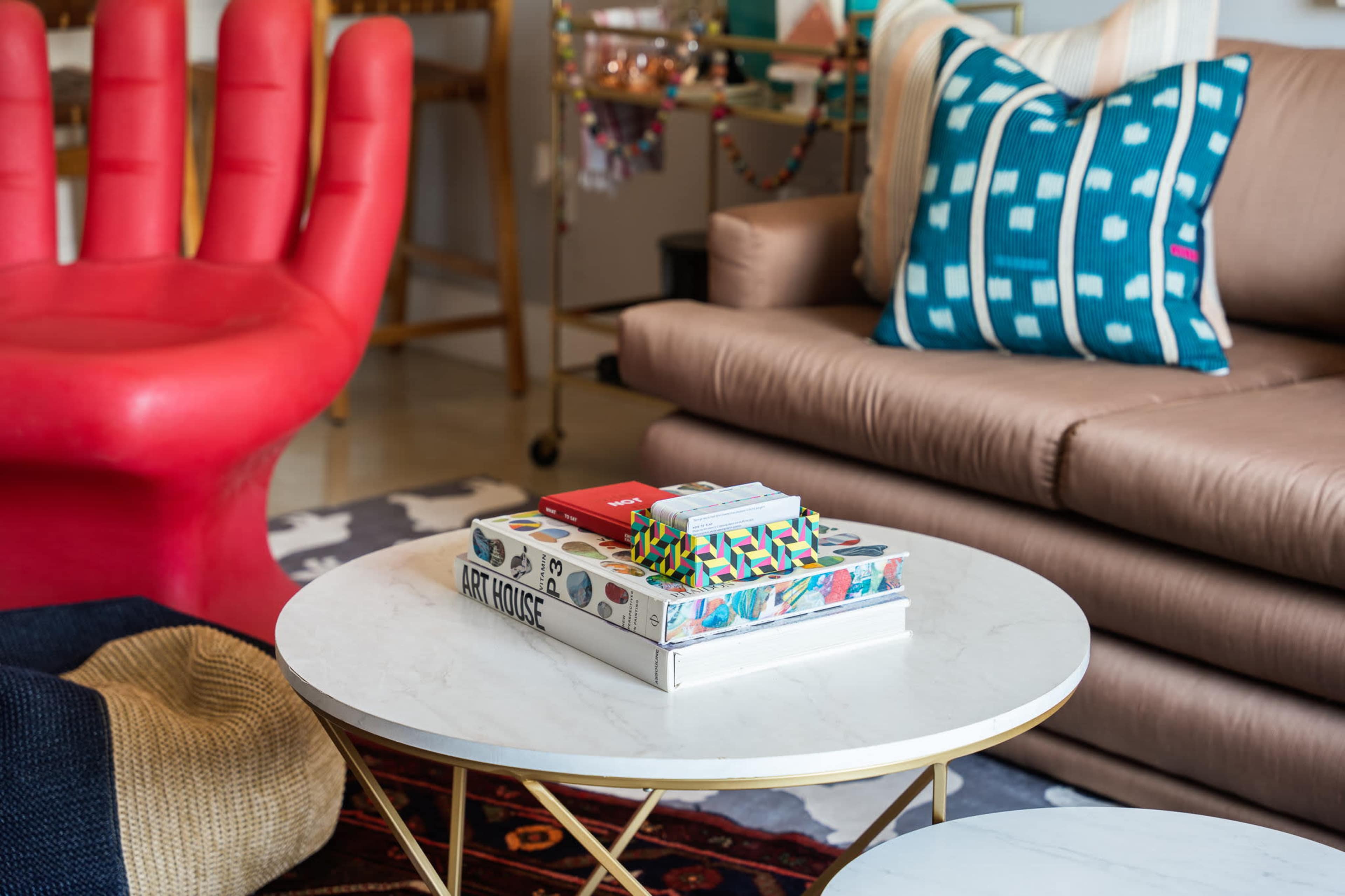 A coffee table with art books, decorative items, and colorful cushions is positioned in front of a beige sofa and a red chair.