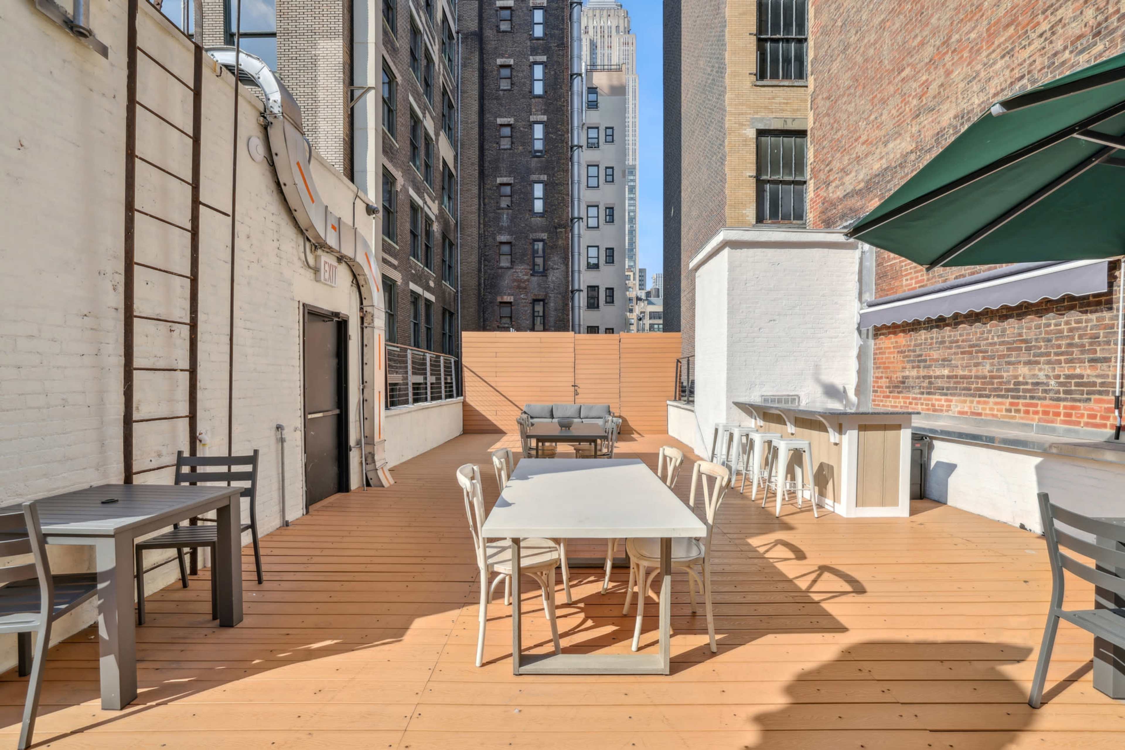 The image shows a spacious rooftop terrace with wooden decking, featuring several tables and chairs, along with a shaded seating area.