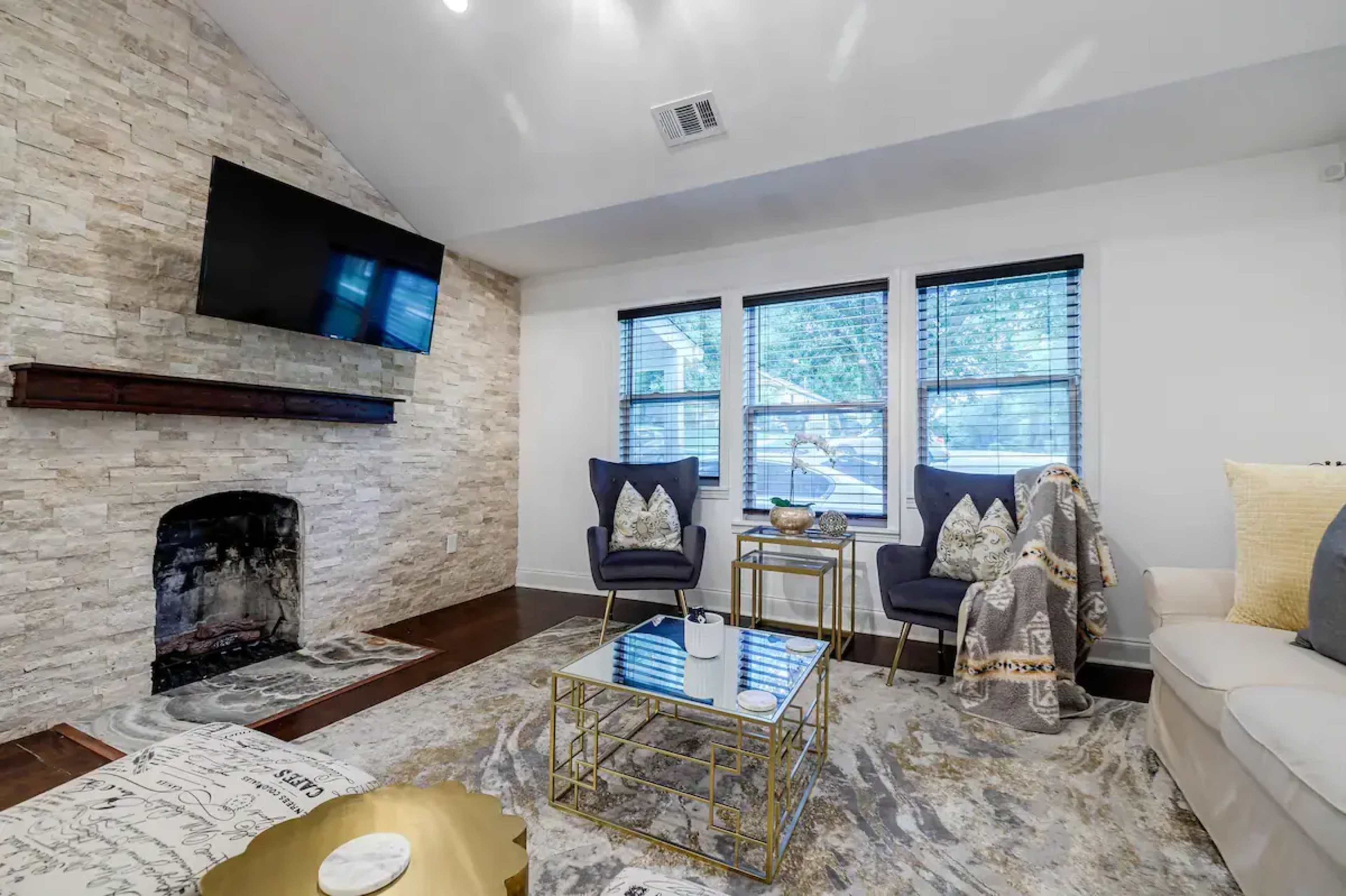 A living room features a stone accent wall with a mounted TV, two armchairs, a coffee table, and large windows allowing natural light to enter.