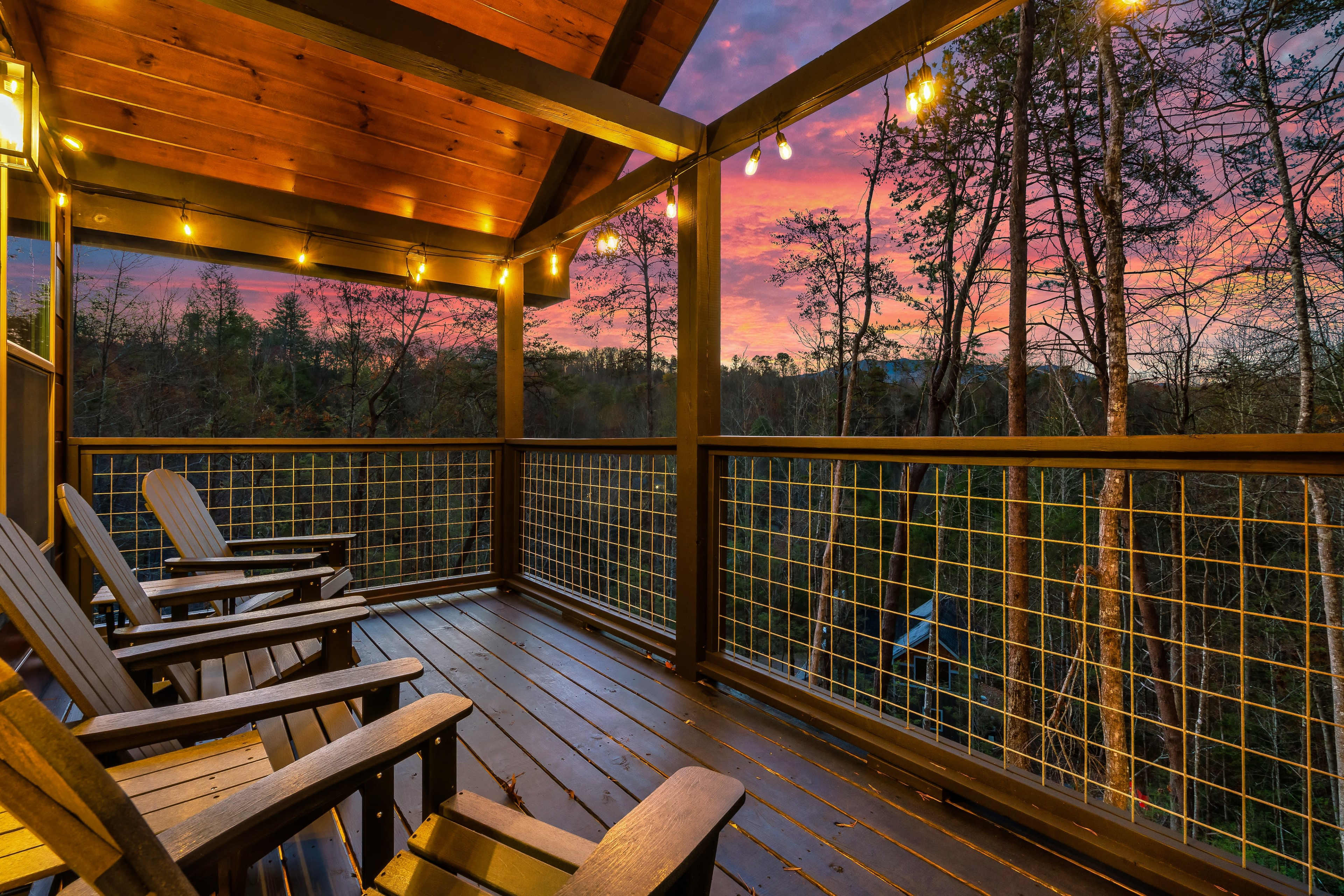 A wooden porch features several lounge chairs overlooking a wooded area under a colorful sunset sky.