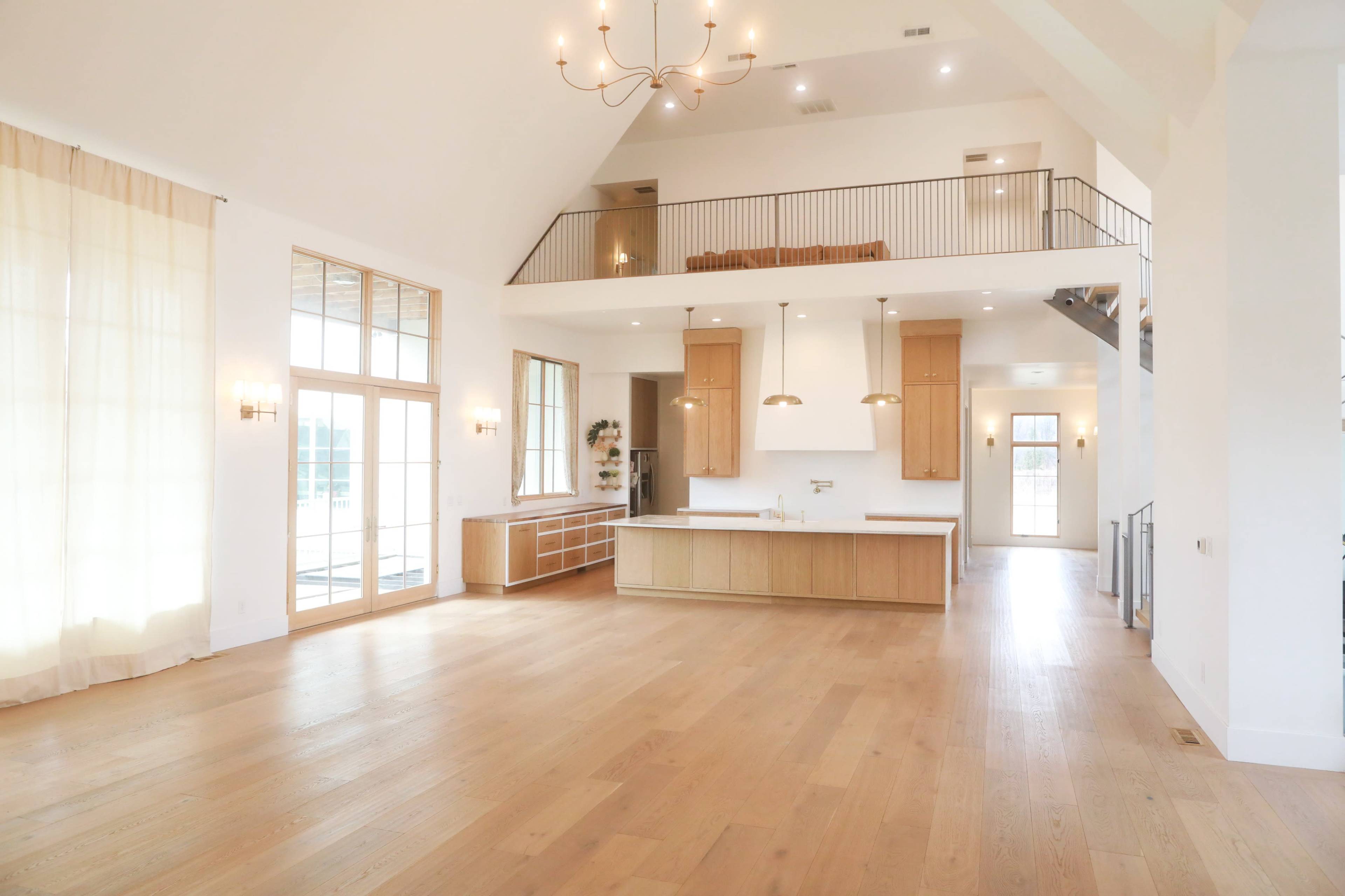 The image shows a spacious, modern open-concept living area with high ceilings, large windows, and a kitchen island at the center.