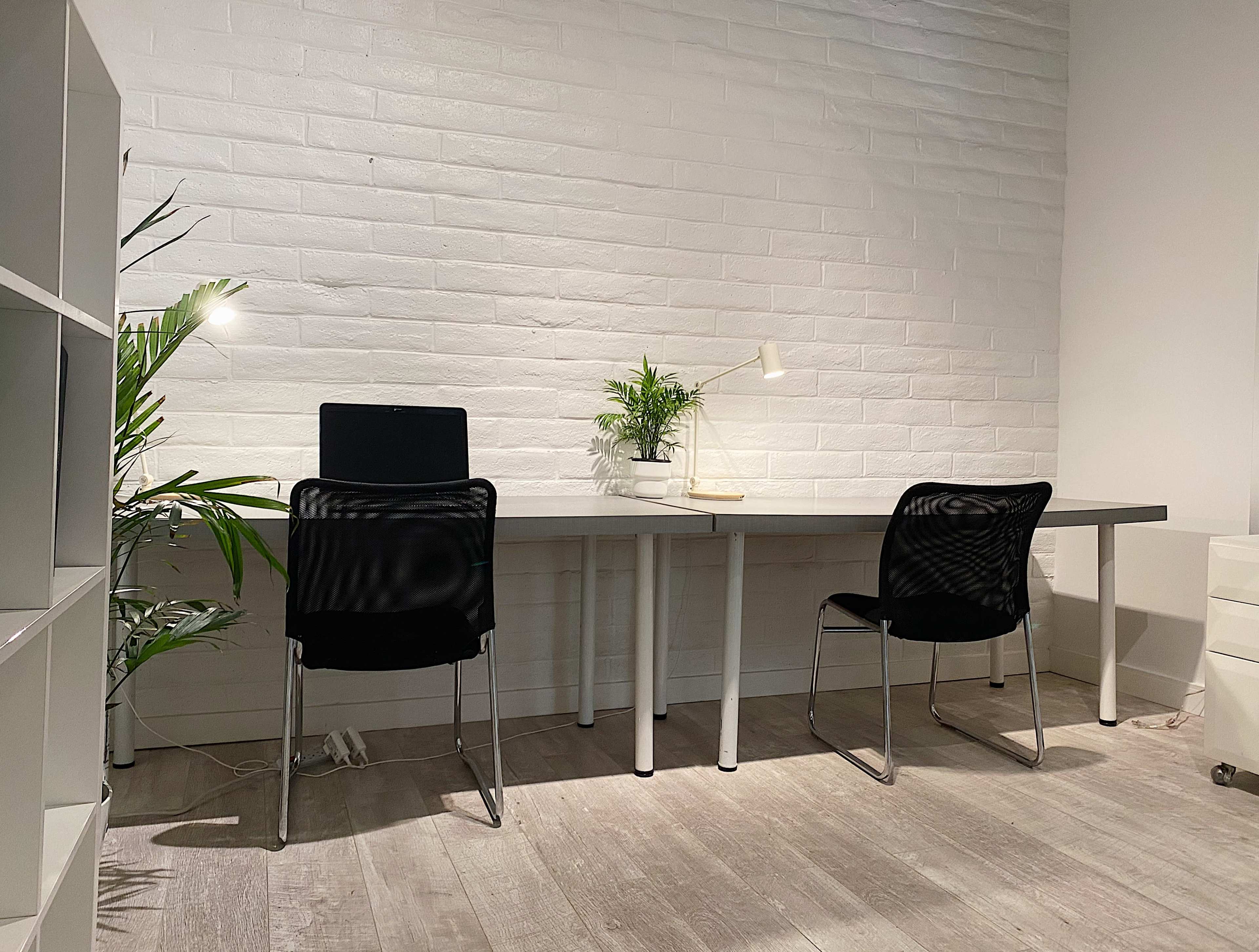 A minimalistic workspace features two black chairs at a light gray table, with a small plant and a lamp on the table against a white brick wall.