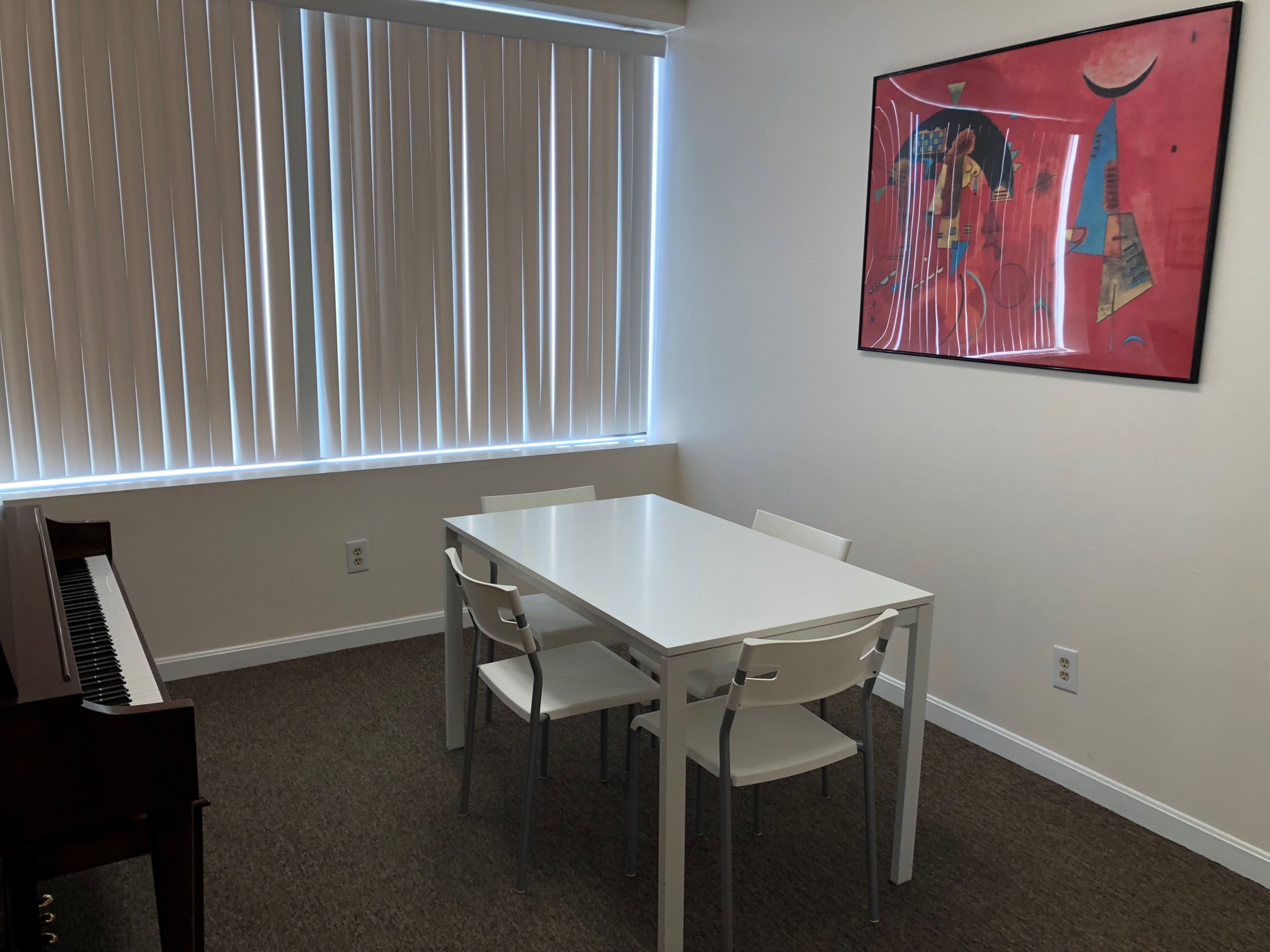 A small, furnished room with a white table and three chairs, a piano against one wall, and a colorful artwork hanging above the table.
