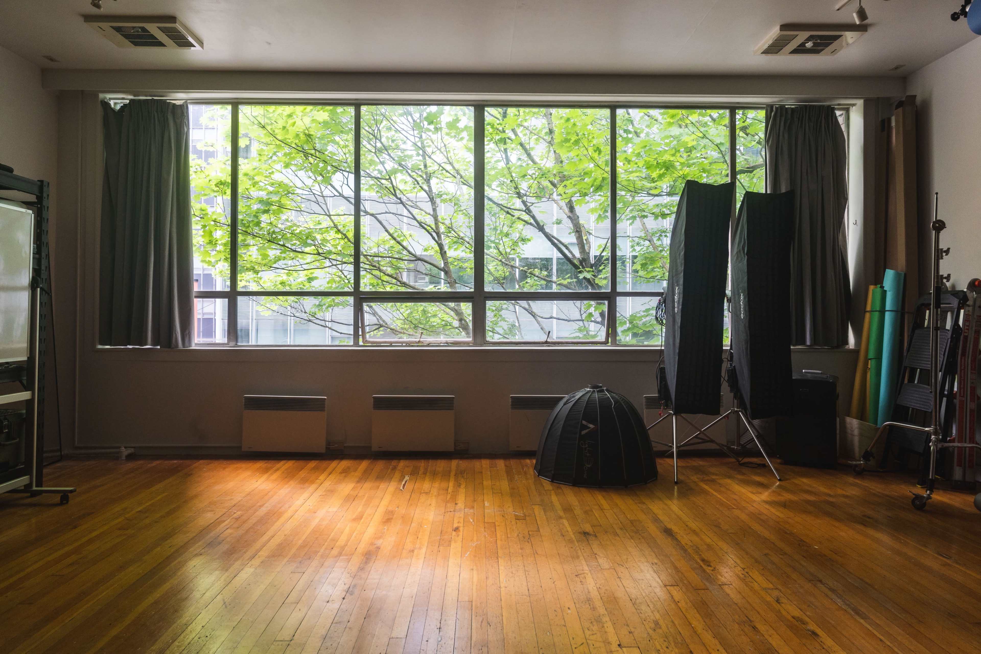 A spacious room with large windows providing natural light, featuring wooden flooring and some studio equipment along the walls.