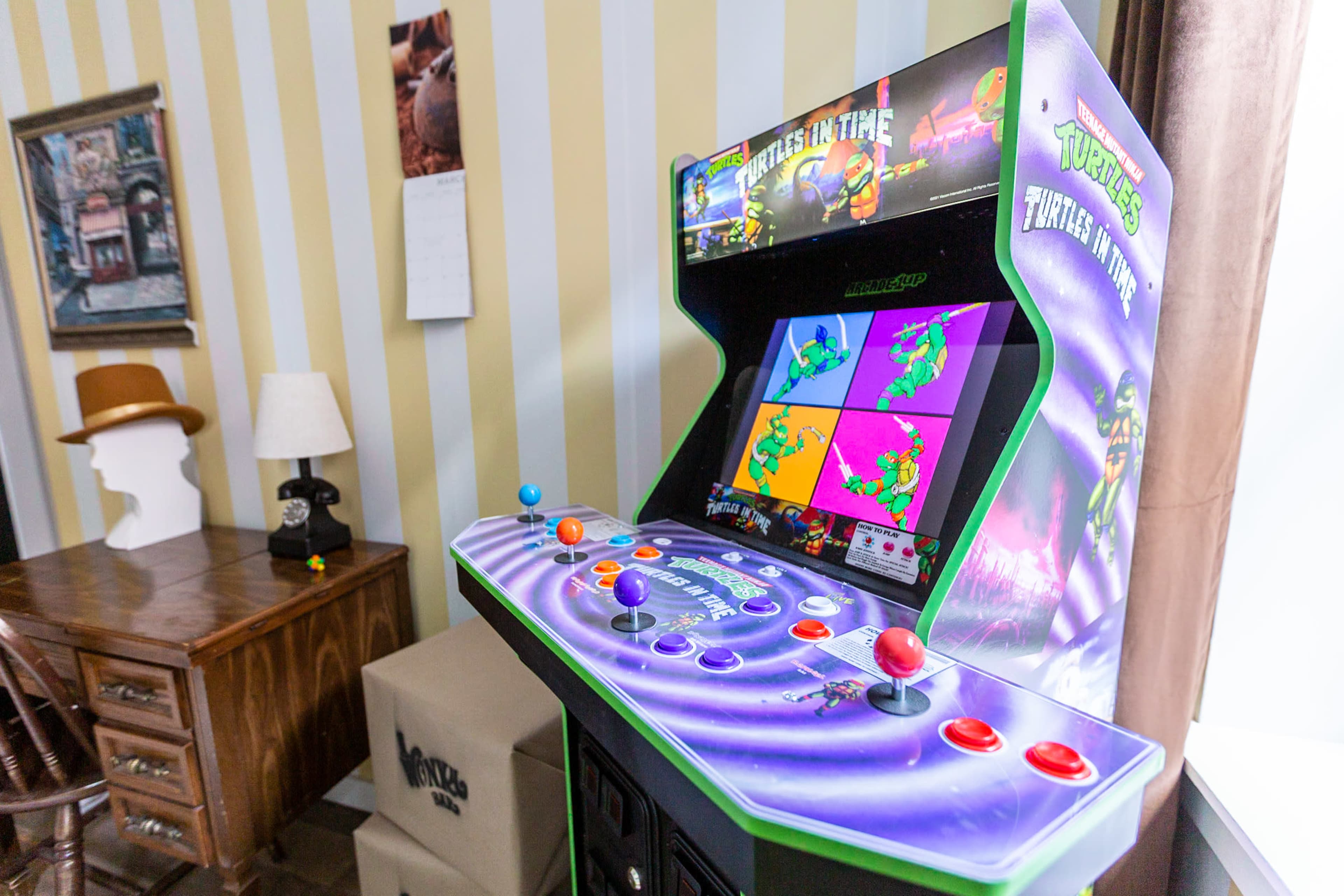A vintage arcade game cabinet featuring "Teenage Mutant Ninja Turtles," set in a room with striped wallpaper, a wooden desk, and various decor items.