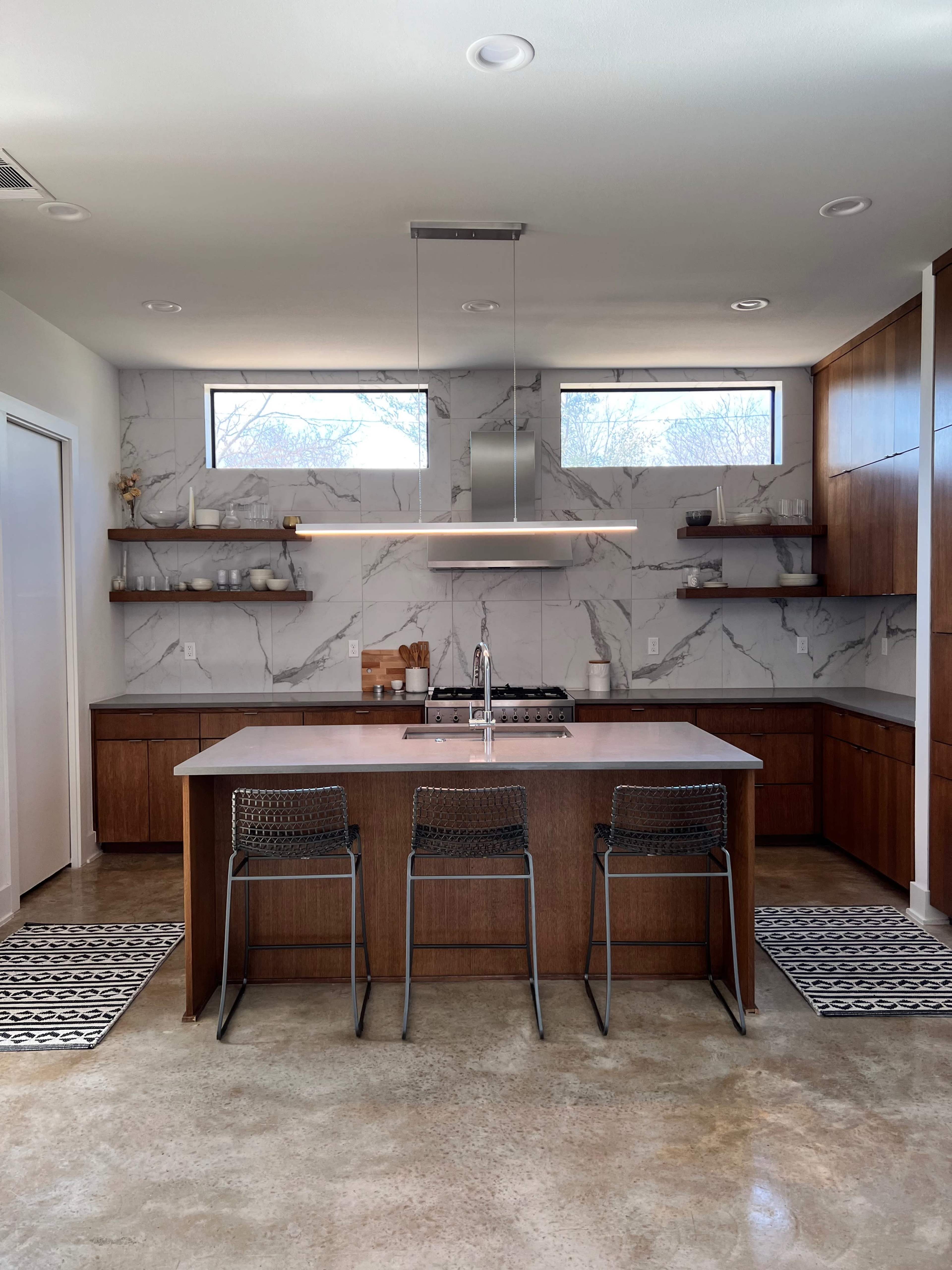 A modern kitchen featuring a central island with three metal chairs, dark wood cabinetry, and marble-patterned backsplash.
