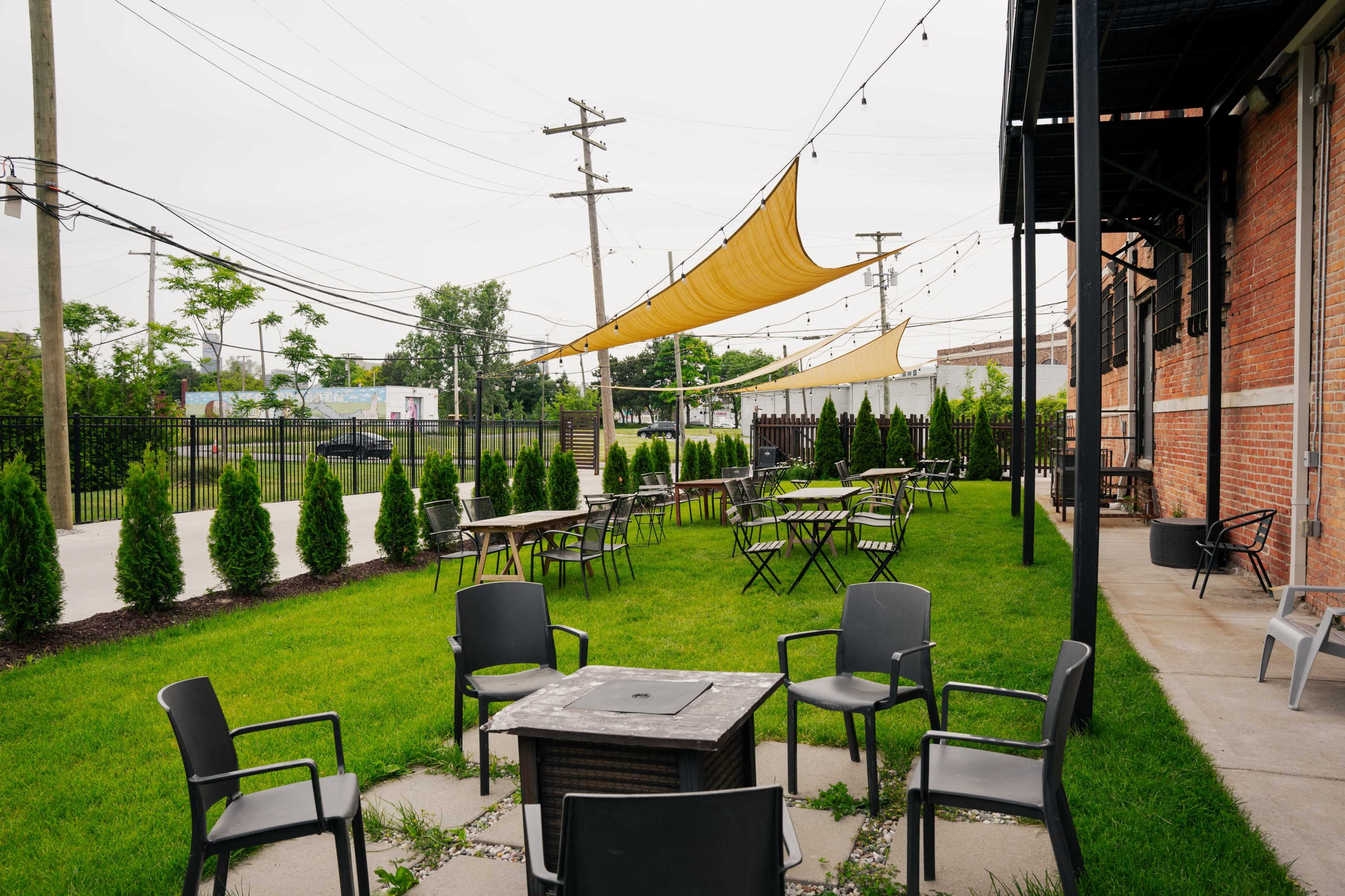 An outdoor seating area with metal chairs arranged around a fire pit, surrounded by neatly trimmed grass and small trees, with fabric canopies overhead.
