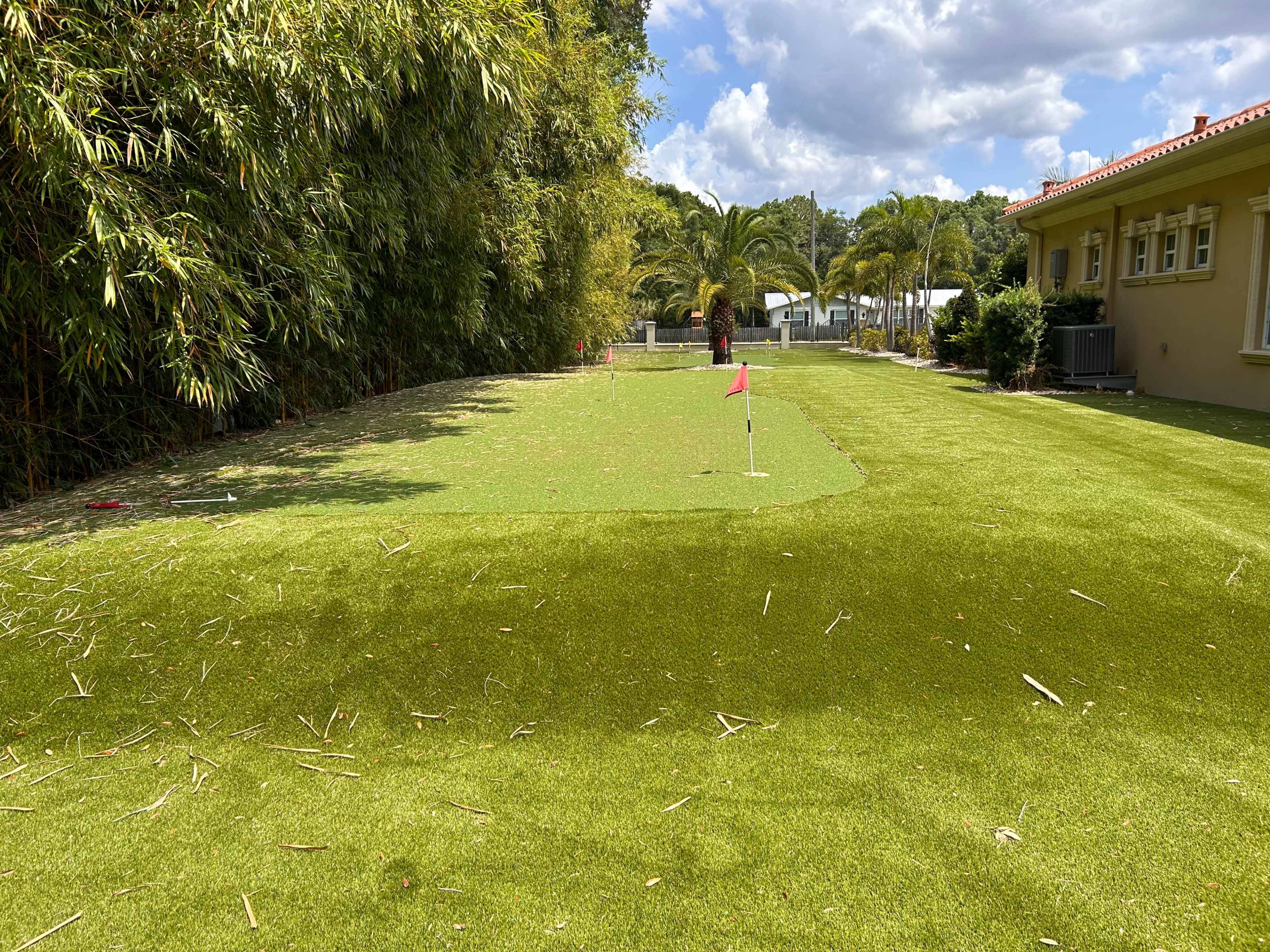 A manicured putting green with a flag is set in a landscaped garden, surrounded by bamboo and palm trees.