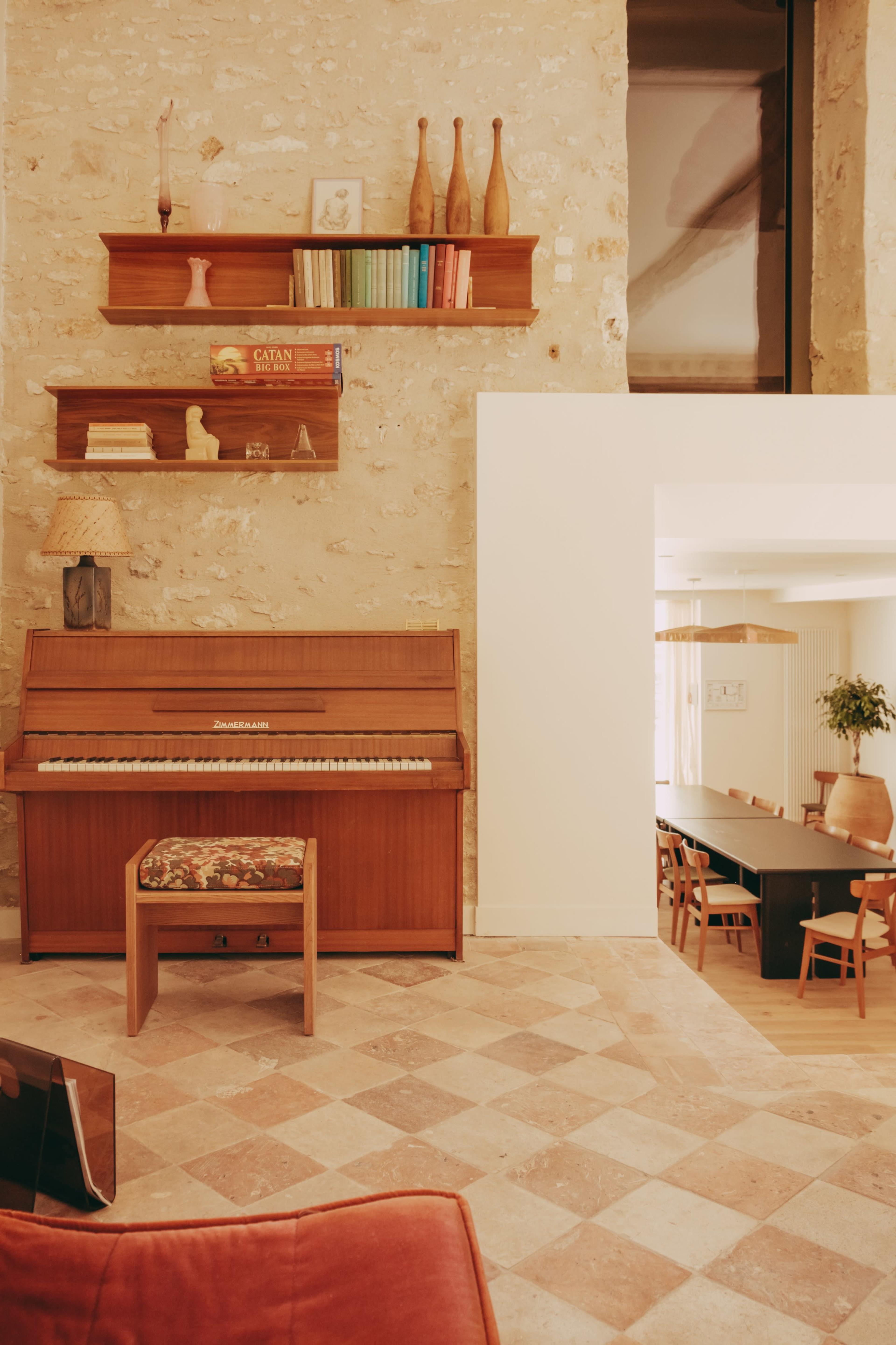 A wooden piano with a floral stool is positioned against a textured stone wall, accompanied by wooden shelves displaying various decorative items.