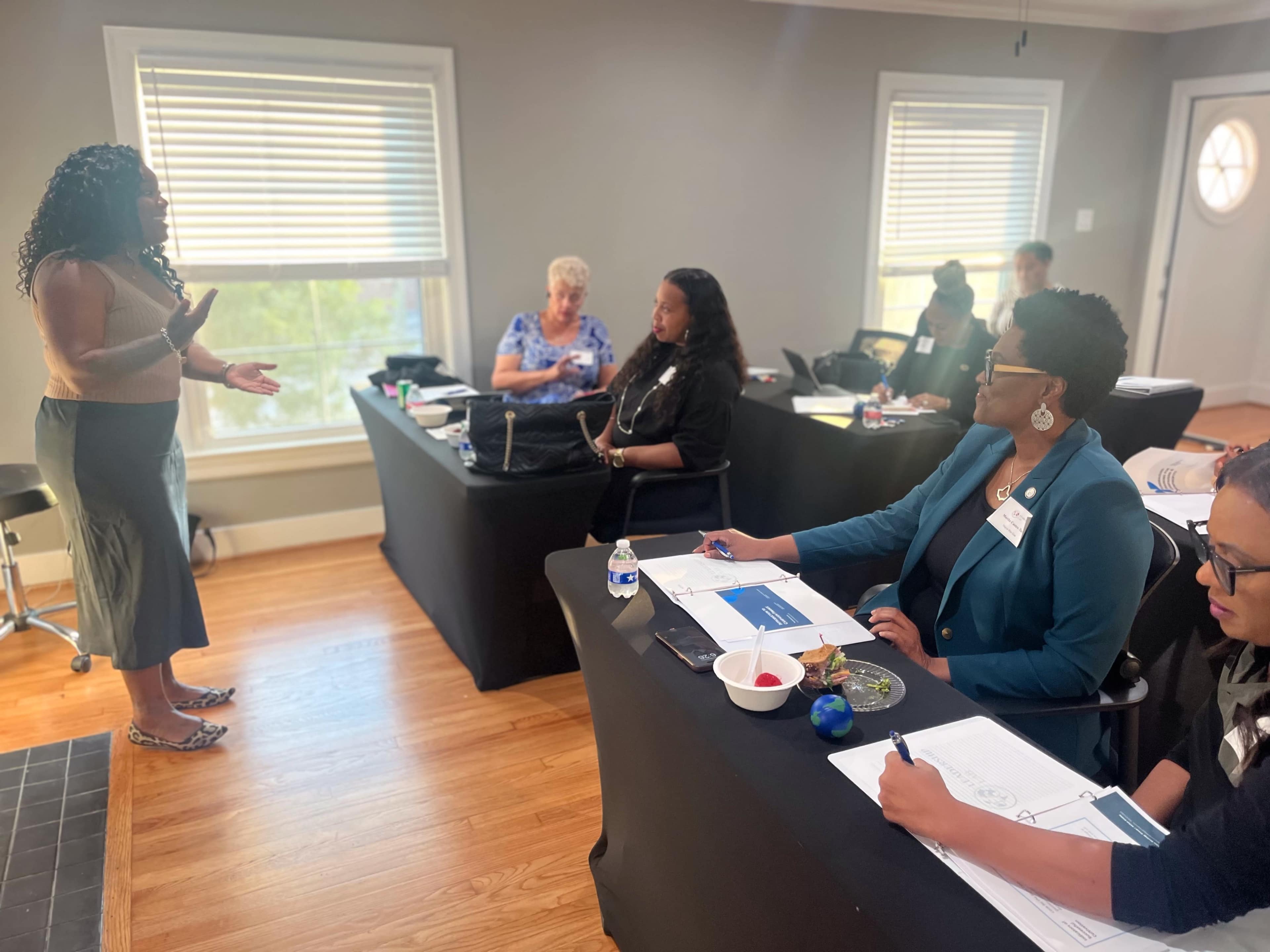 A group of women sit at tables in a classroom setting, while one woman stands and speaks to the audience.