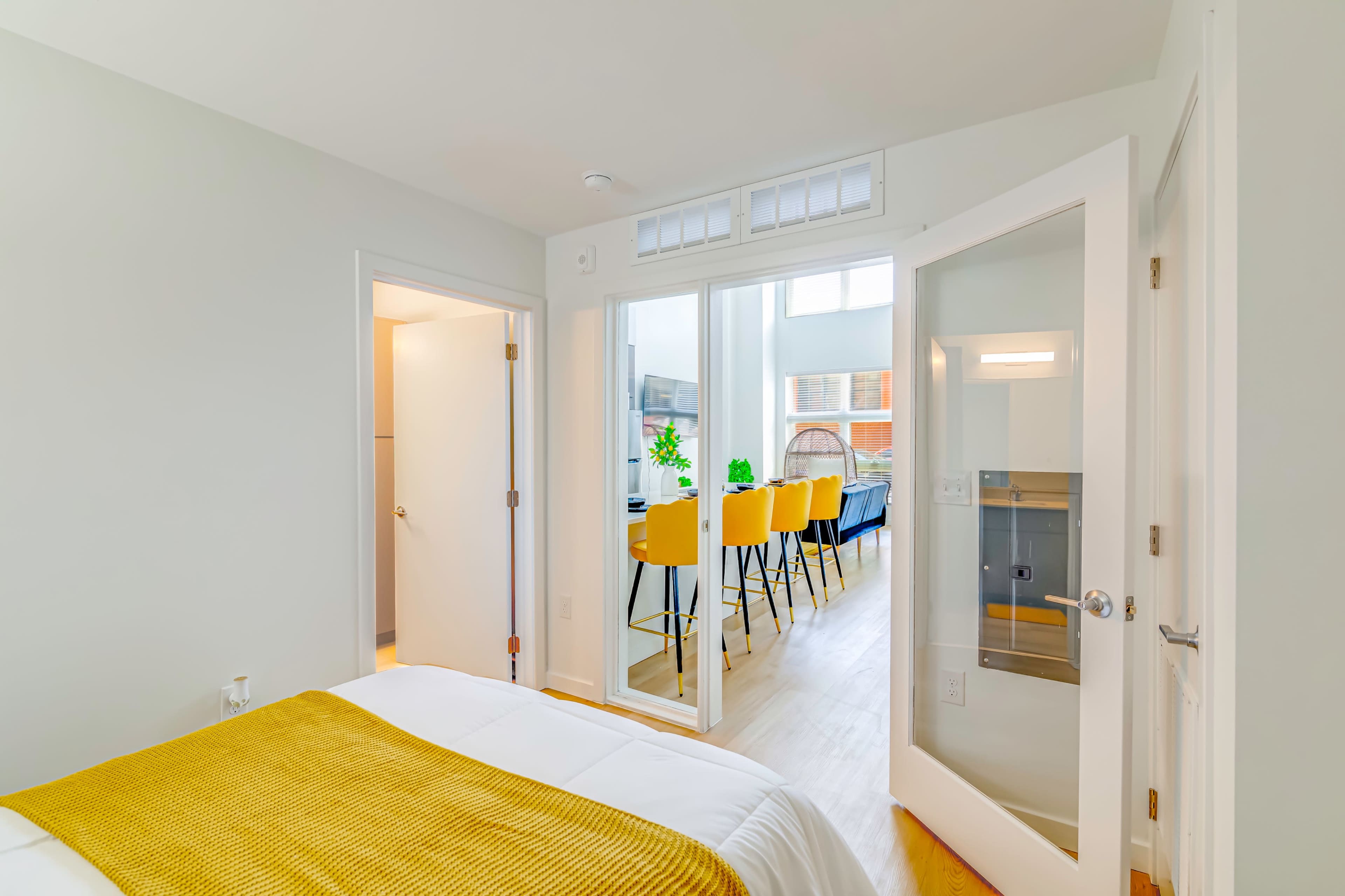 A well-lit bedroom features a white bed with a yellow blanket, leading to an open area with a dining table and chairs visible through glass doors.