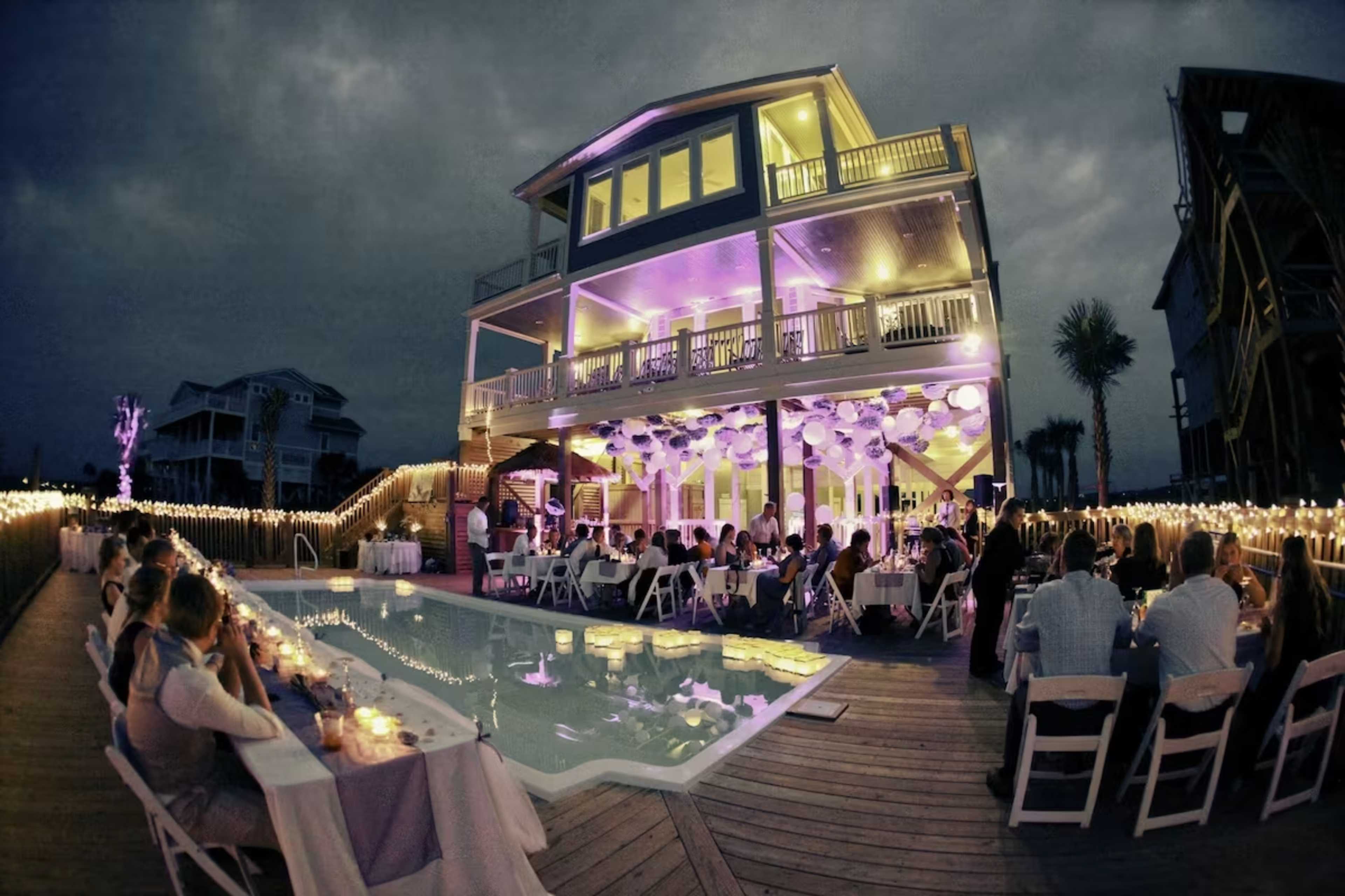 A large gathering is taking place around a pool at a beautifully lit house, decorated with string lights and purple accents, as guests sit at tables.
