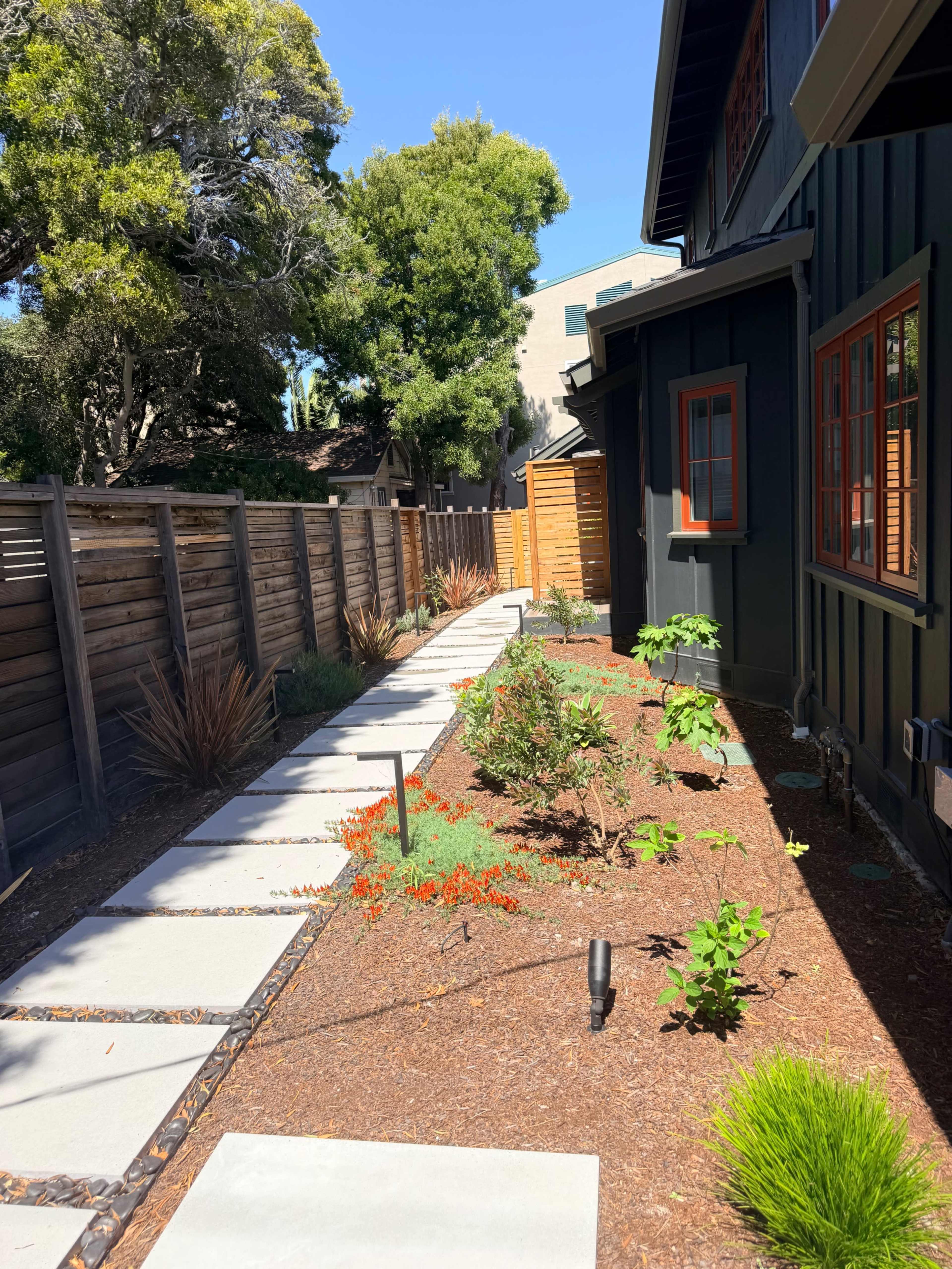 The image shows a walkway lined with concrete stepping stones leading through a garden with various plants alongside a house and wooden fence.