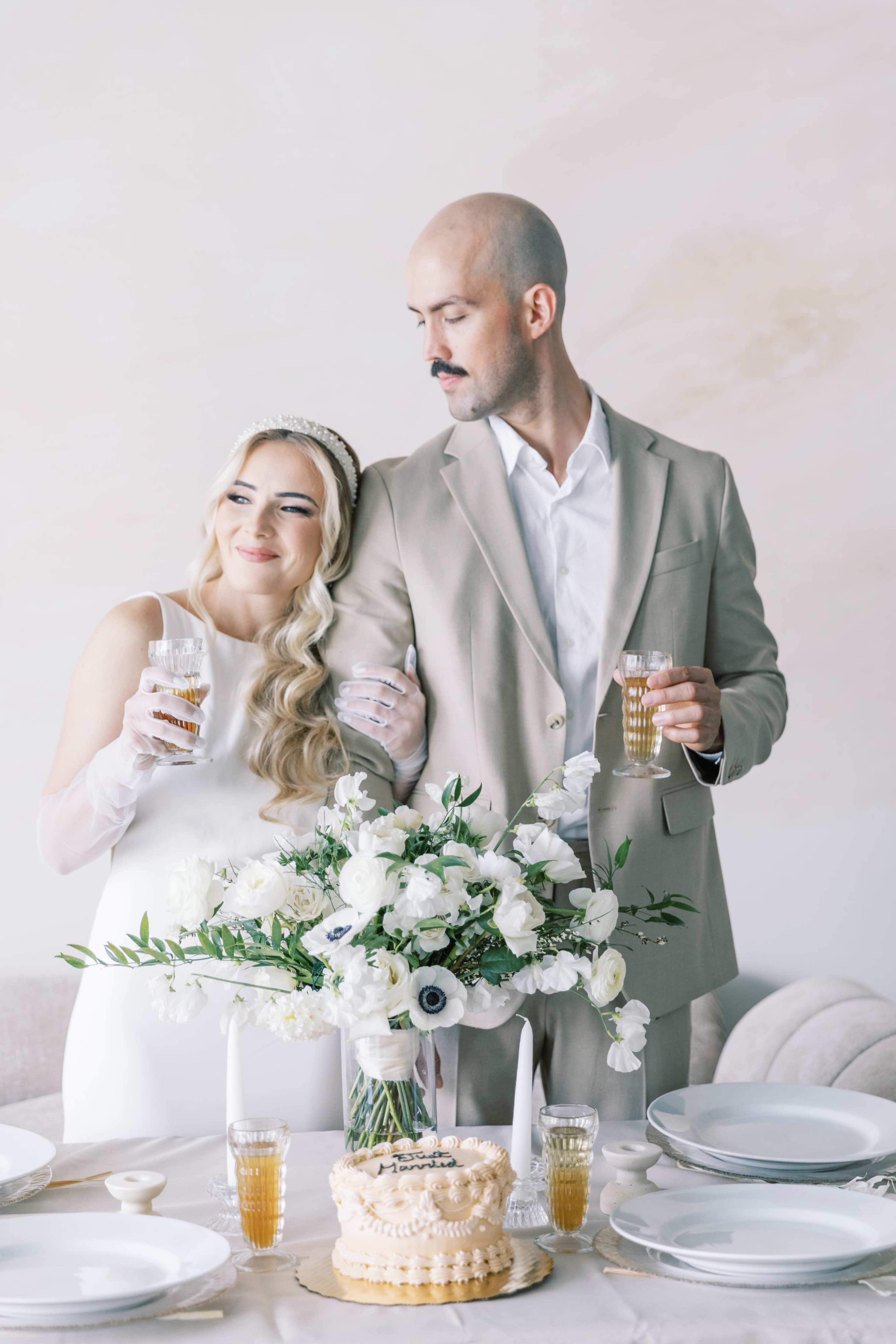 A couple stands together at a elegantly set table, holding drinks and surrounded by flowers and a cake.