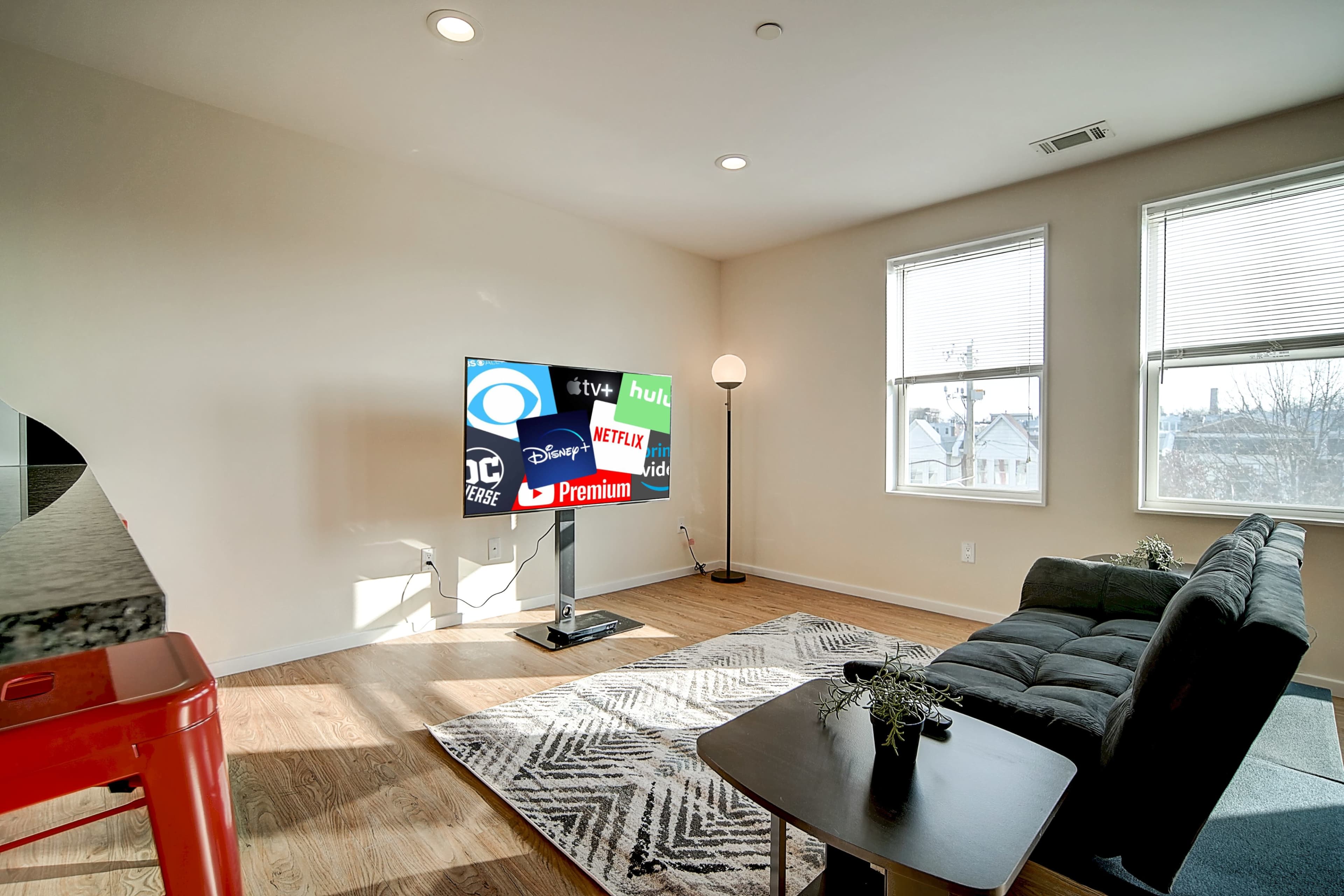 A living room features a television displaying various streaming service logos, with a couch and a lamp situated beside large windows.