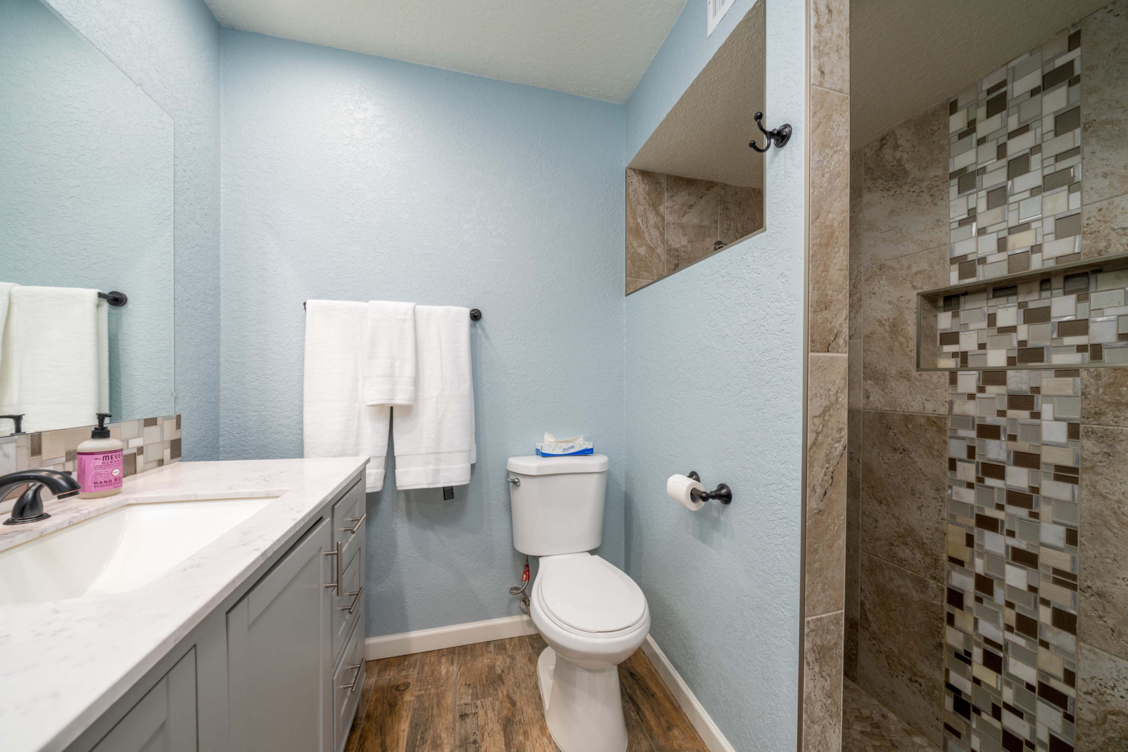 A clean bathroom features a light blue wall, a white vanity with a sink, a toilet, and a tiled shower area.