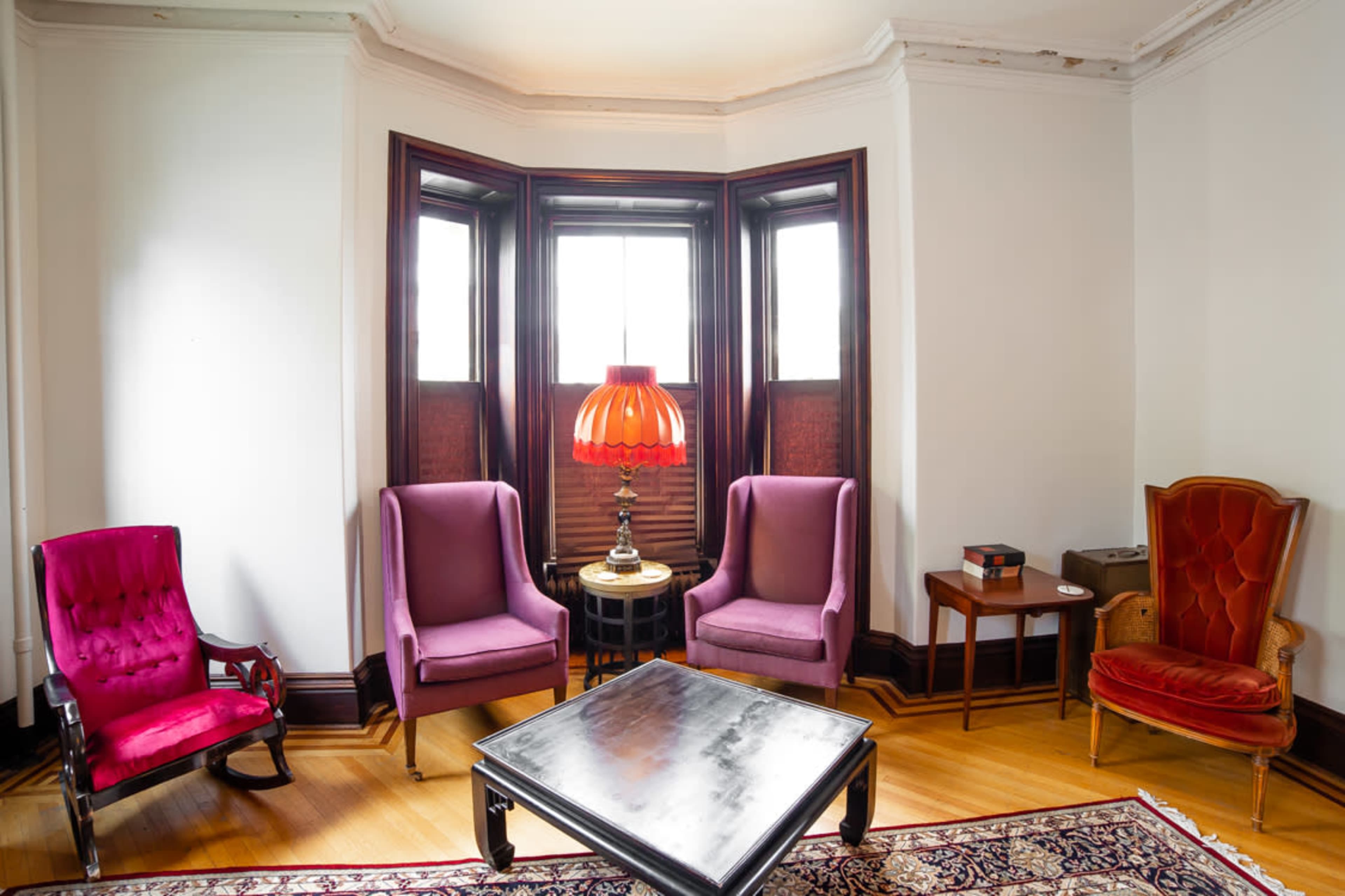 A living room features three upholstered chairs and a table with a lamp near a bay window, surrounded by wood paneling and a patterned rug.