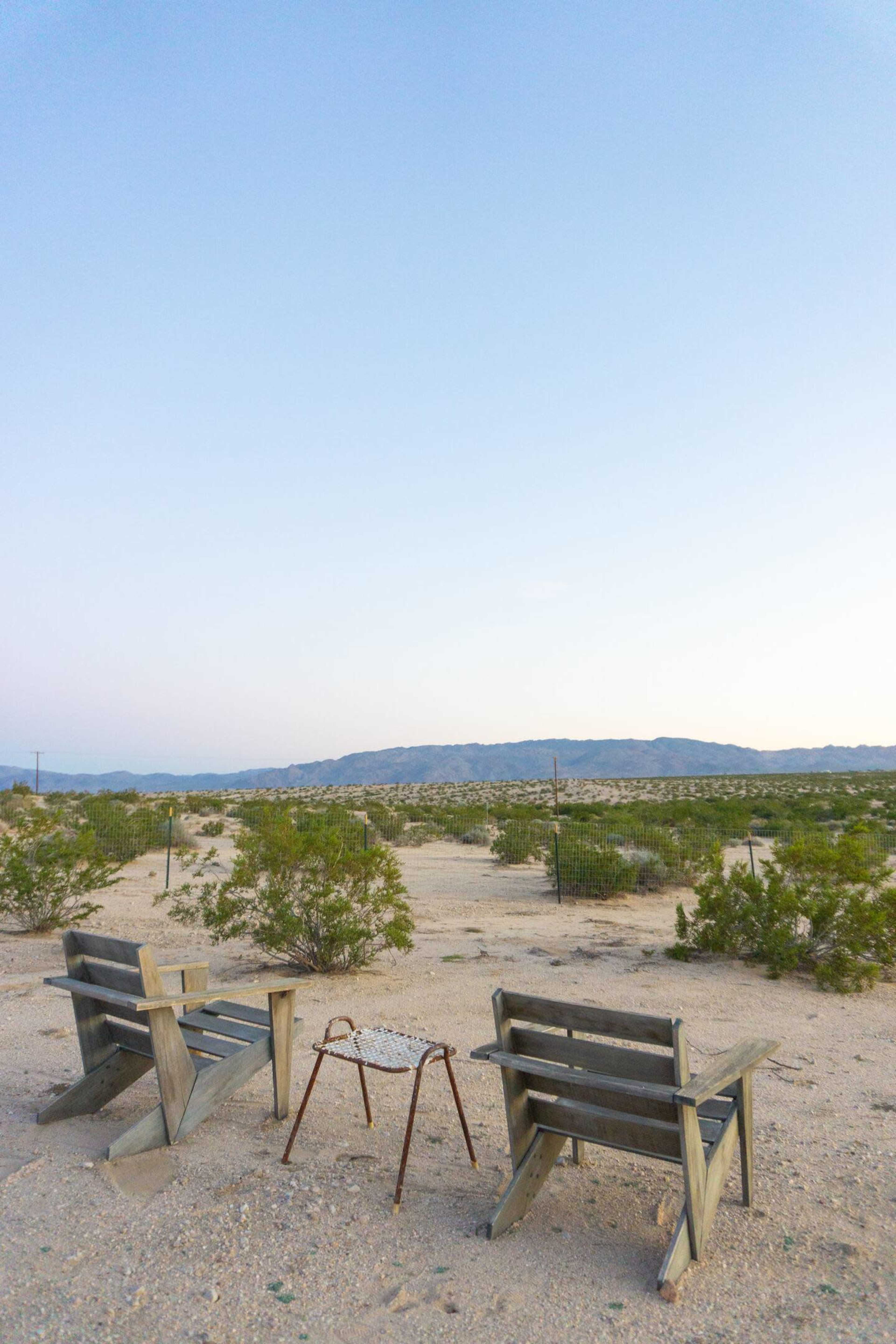 Two wooden chairs and a small table sit in a barren desert landscape under a clear sky.