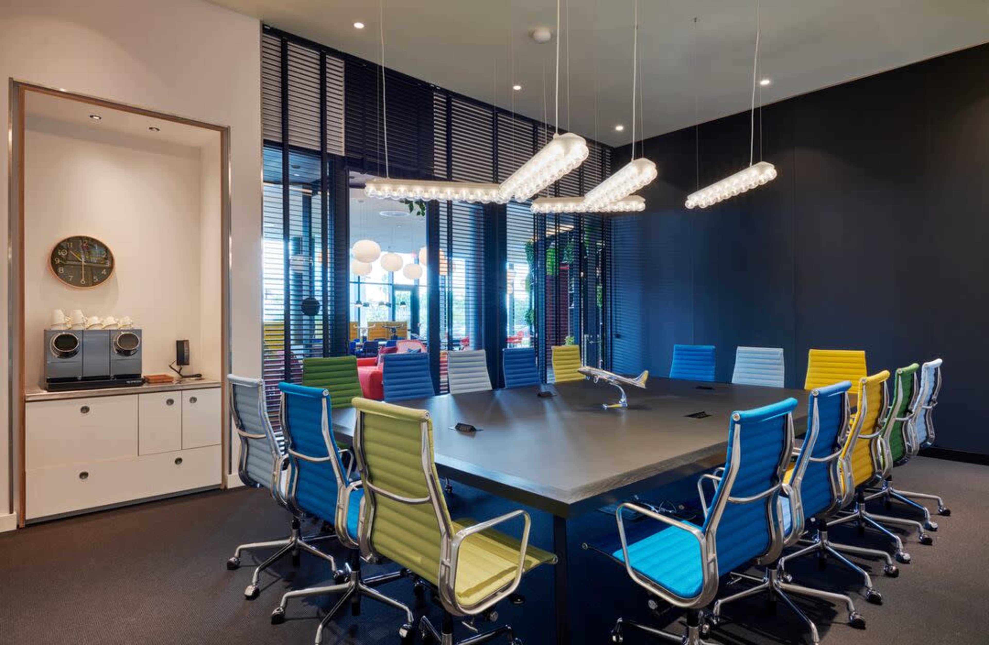 A modern conference room with a long table surrounded by colorful chairs and large windows allowing natural light.
