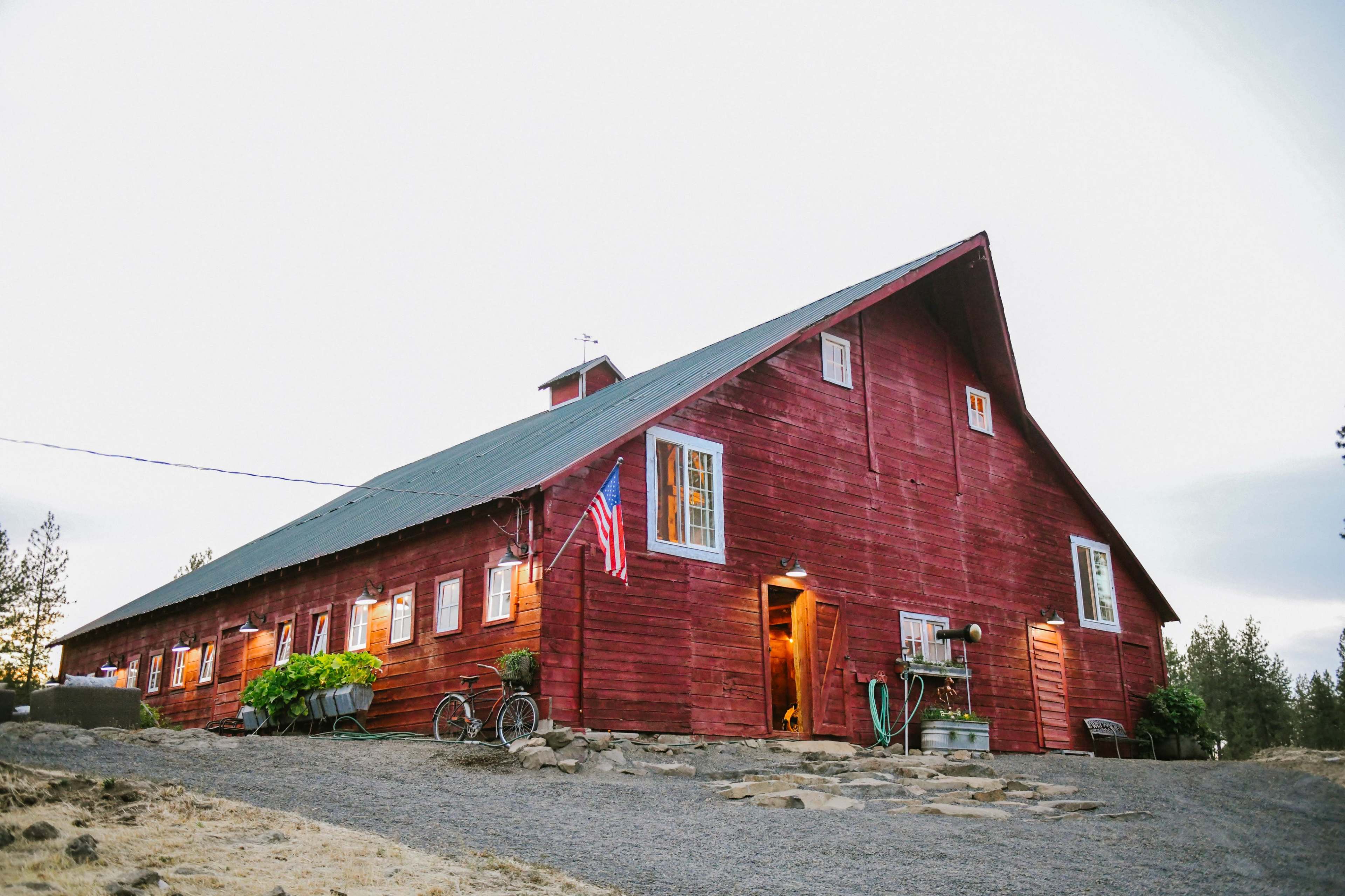 A large red barn with a sloped roof stands on a gravel path, featuring windows that emit warm light and an American flag displayed on the side.