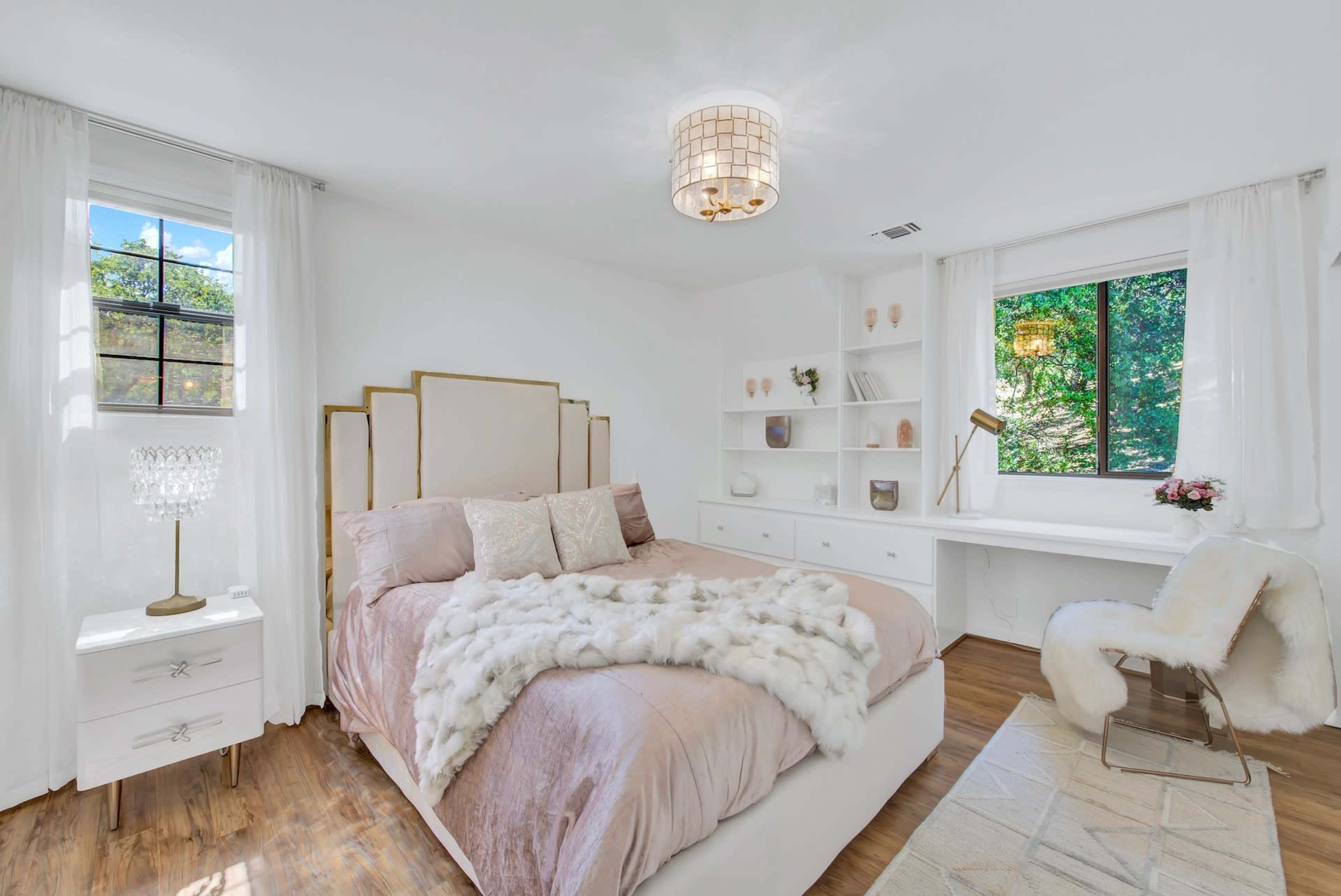 A well-lit bedroom features a bed with a light pink duvet and pillows, a white desk, and a window with sheer curtains overlooking greenery.