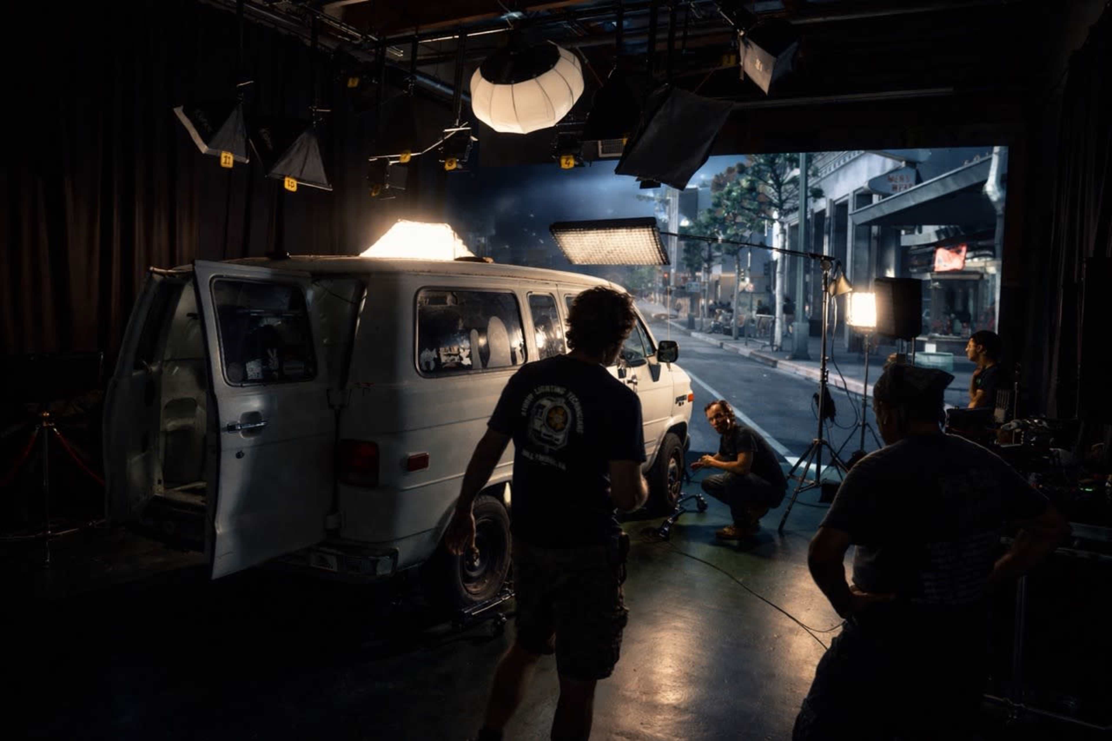 A film crew works on a set featuring a white van, surrounded by studio lights and equipment.