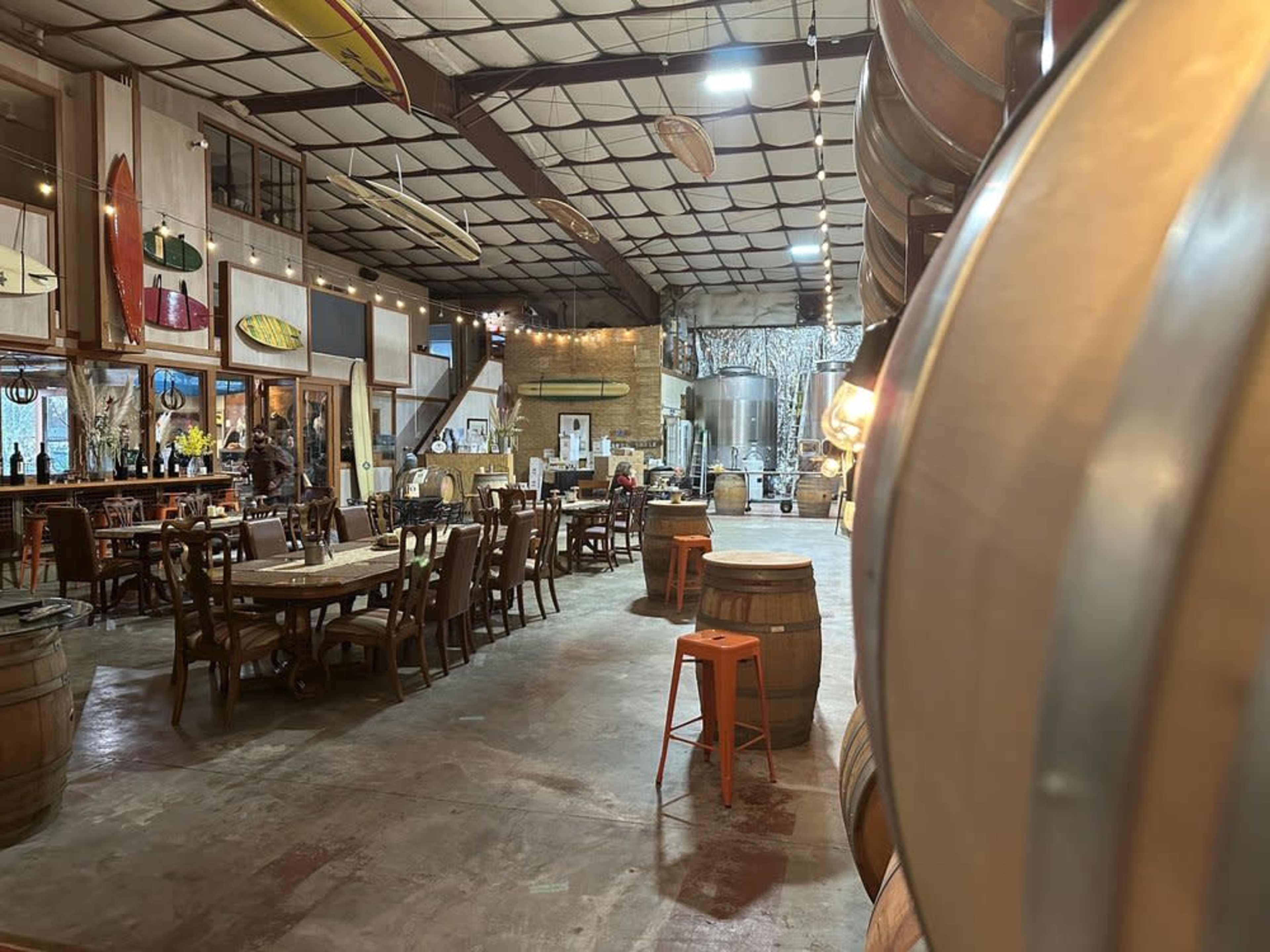The image shows a spacious interior of a rustic venue with wooden tables, barrels, and brewing equipment in the background.