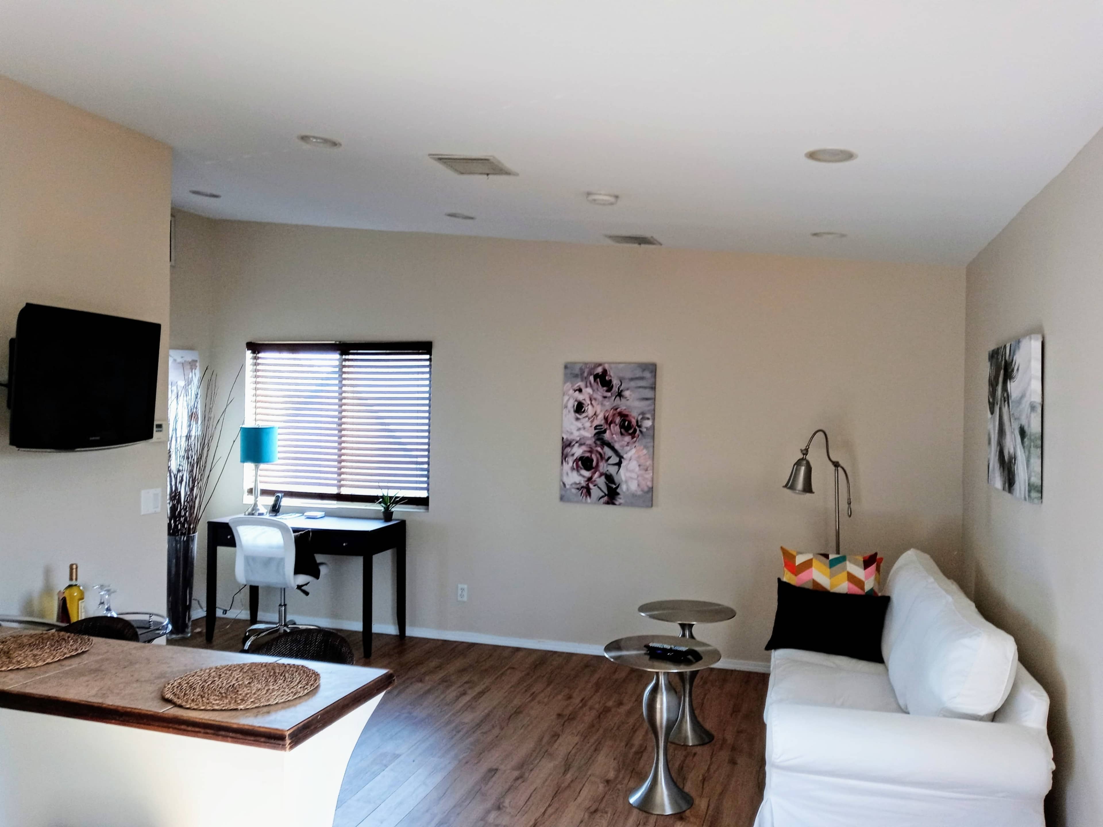 The room features a living area with a white sofa, two side tables, a desk with a chair, and wall art, illuminated by natural light from a window.