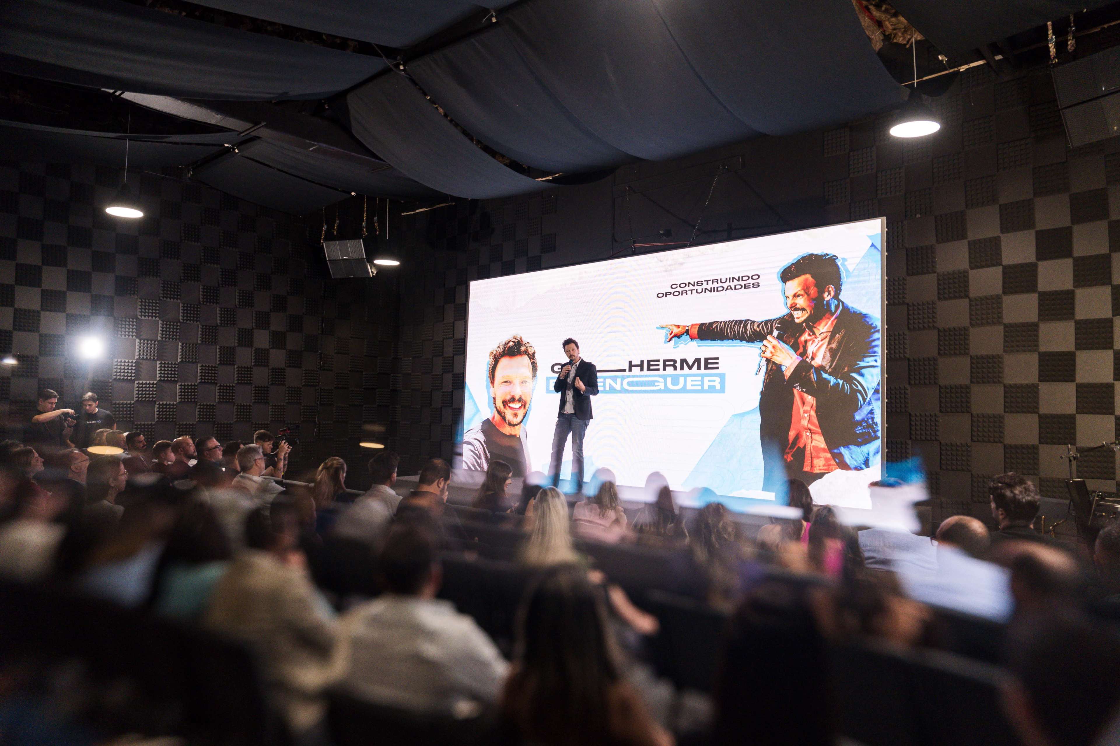 A speaker stands on stage in front of a large screen while an audience listens attentively in a modern event space.