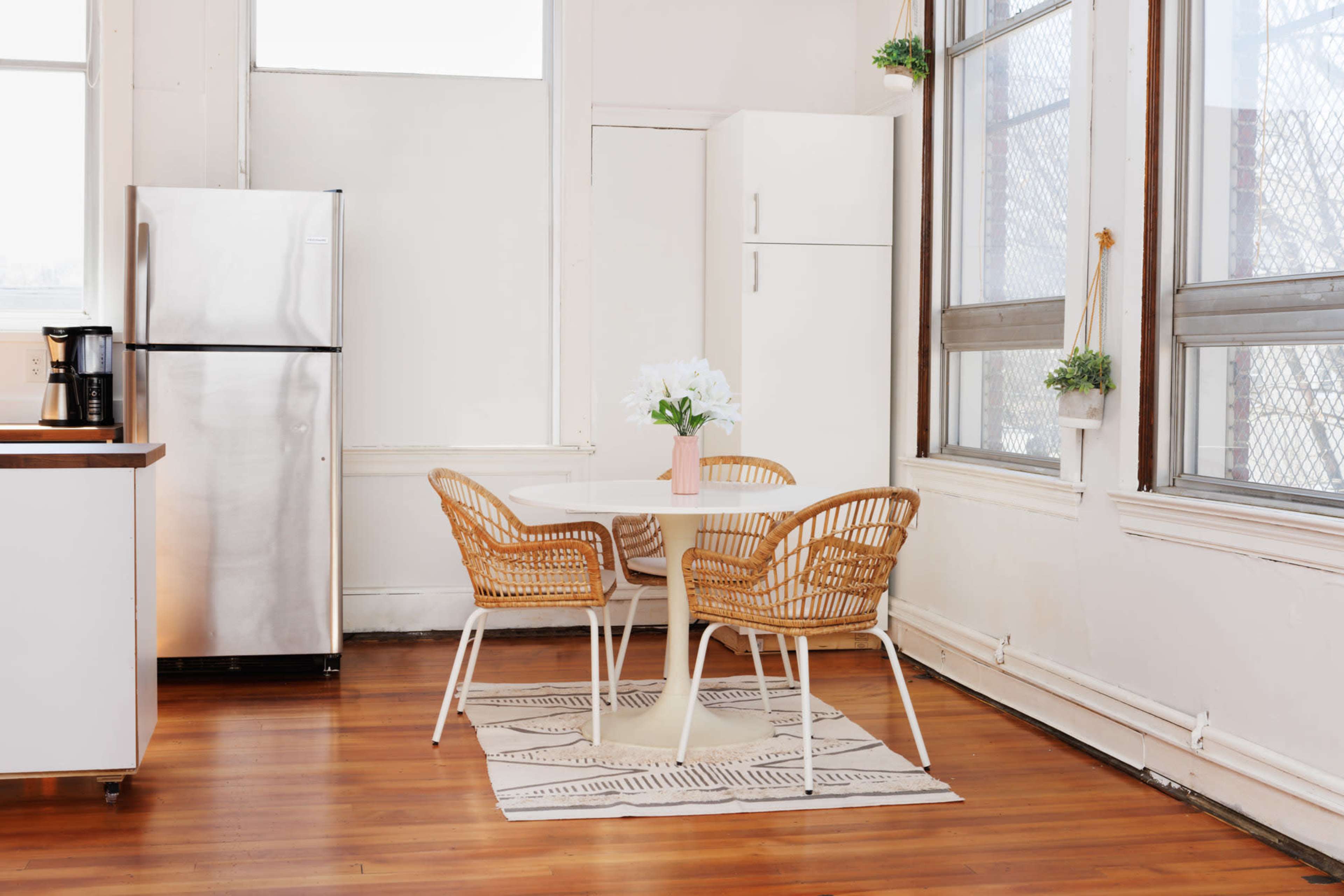 A kitchen area features a round table with two woven chairs, a stainless steel refrigerator, and large windows allowing natural light to enter.