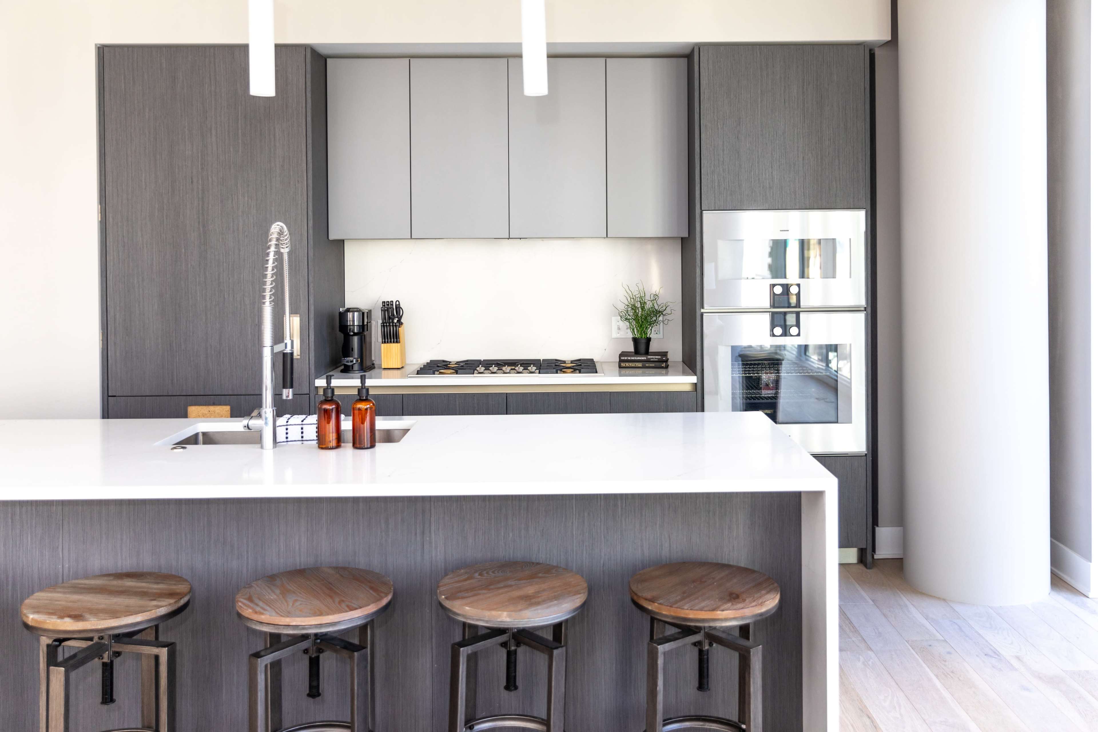 A modern kitchen featuring a gray cabinetry, a white countertop, a gas stove, and four wooden bar stools arranged at a kitchen island.