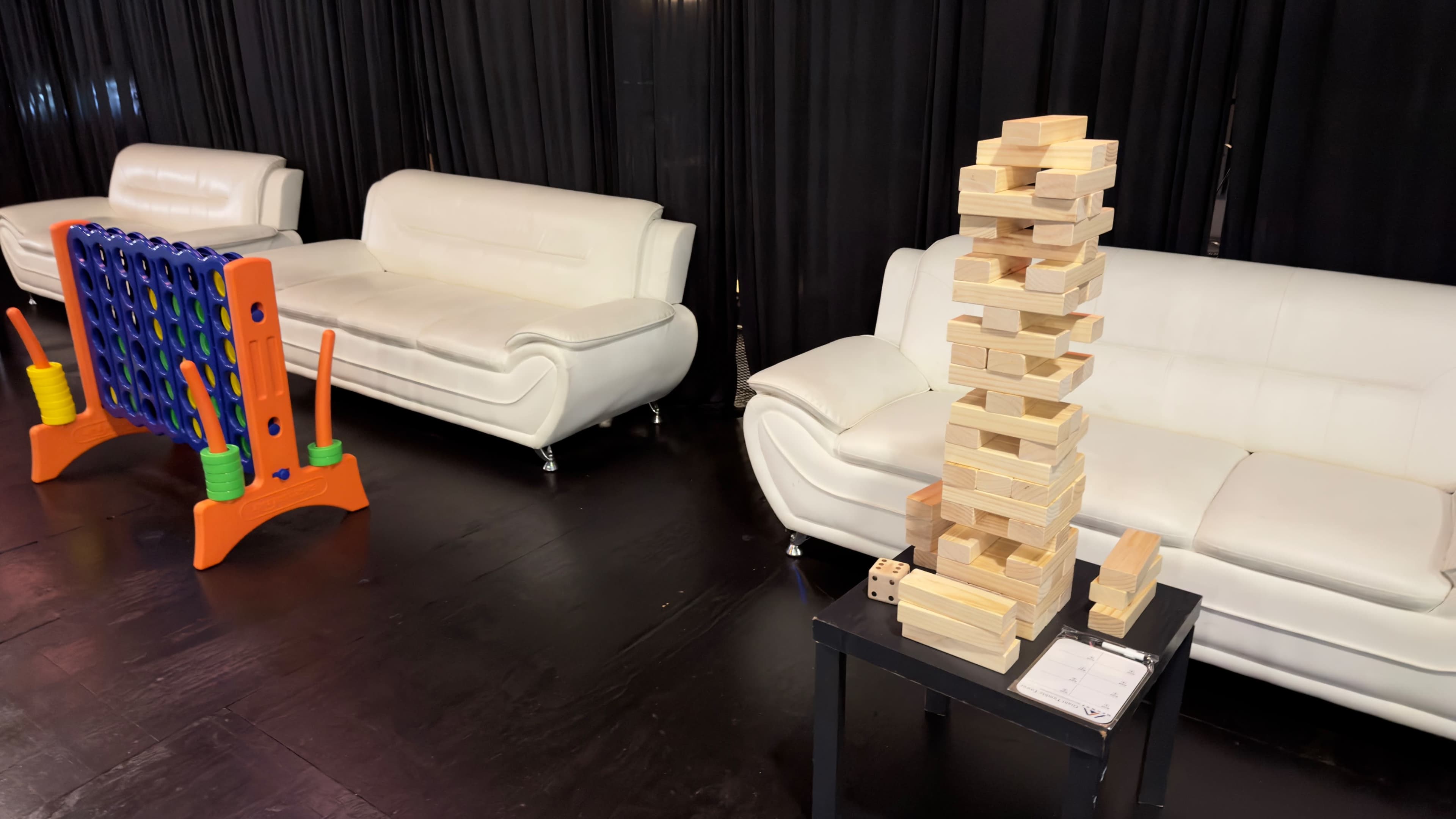The image shows two white sofas facing a tall stack of wooden blocks on a black table, with a colorful game structure behind them.