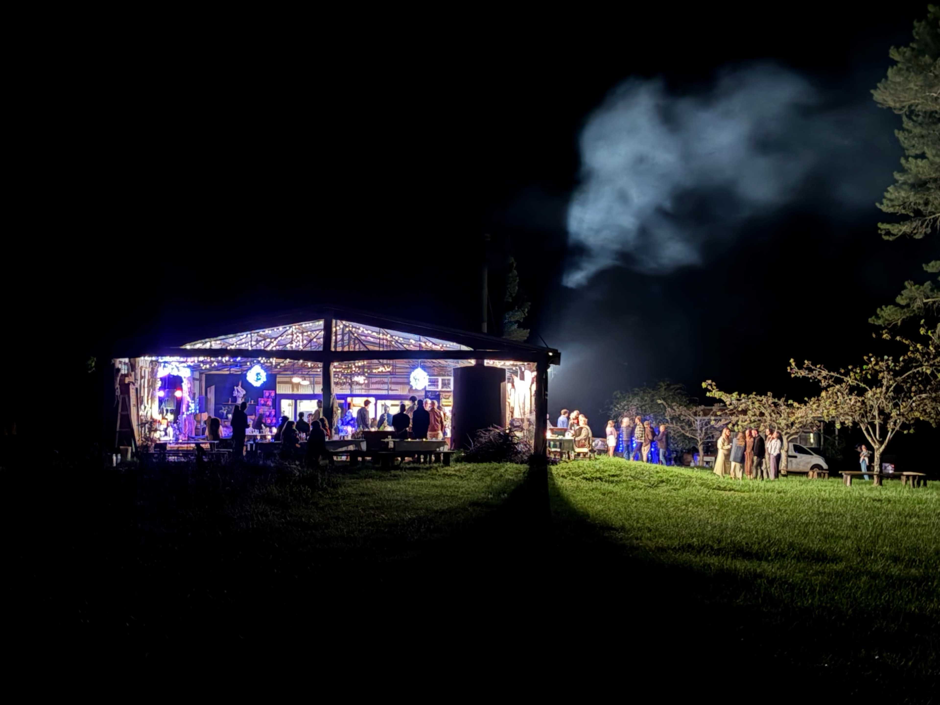 A lively gathering takes place outside a well-lit wooden structure at night, with guests mingling and smoke rising in the background.