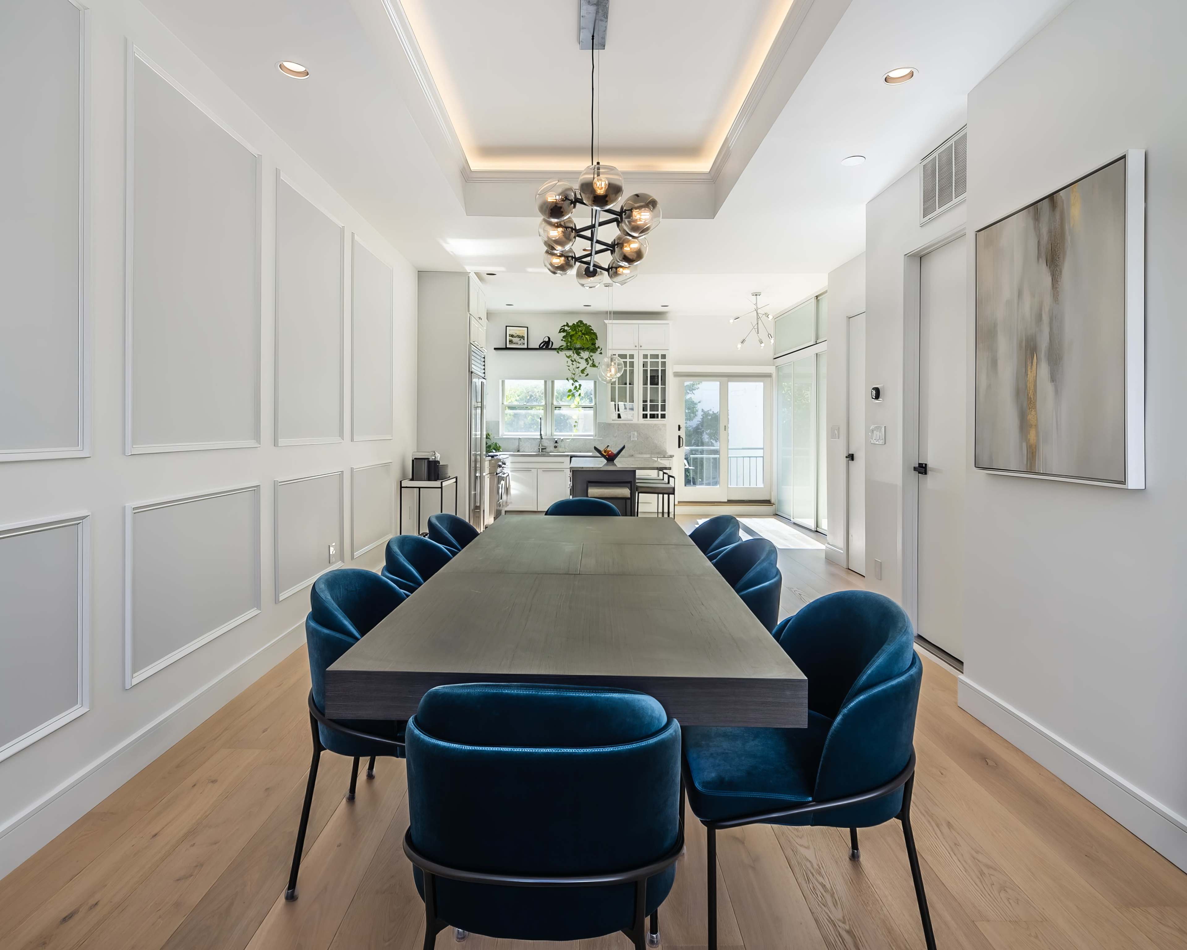 A modern dining room features a long rectangular table surrounded by blue upholstered chairs, with a chandelier above and a kitchen visible in the background.