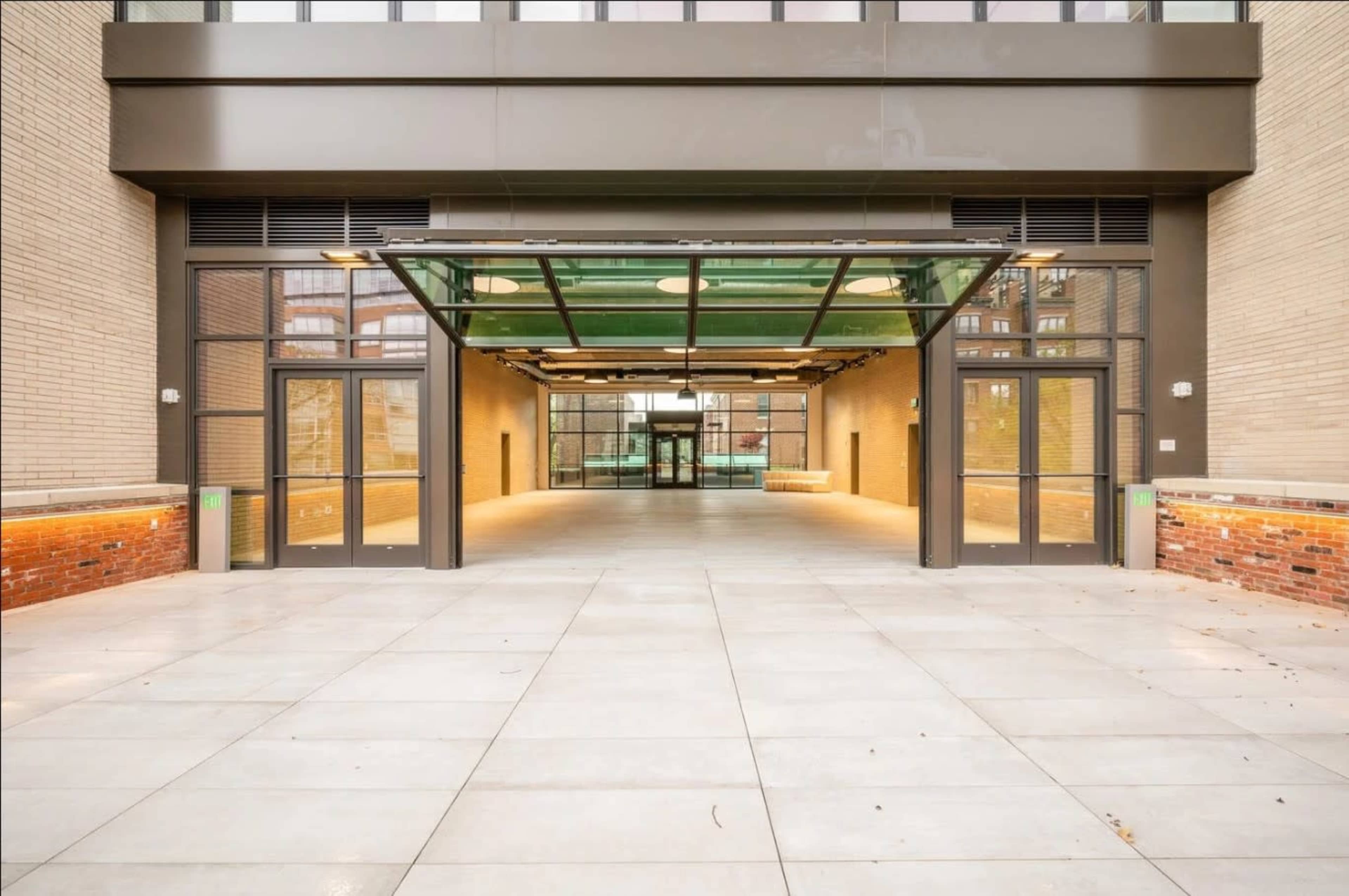 The image shows a large, open entrance with two glass doors leading into a well-lit interior space featuring a flat, tiled floor and exposed brick walls.