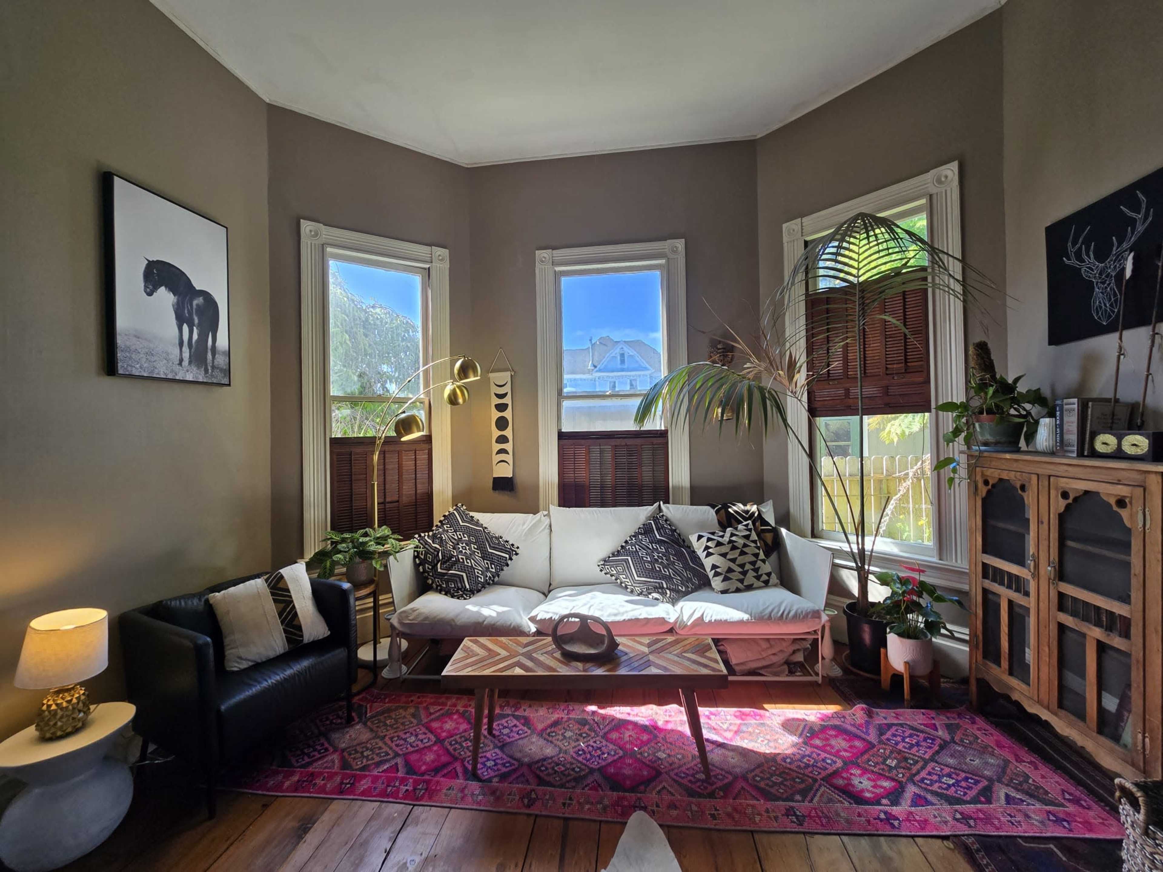 A cozy living room features a white sofa with patterned pillows, a black armchair, a wooden coffee table on a colorful rug, and large windows allowing natural light.