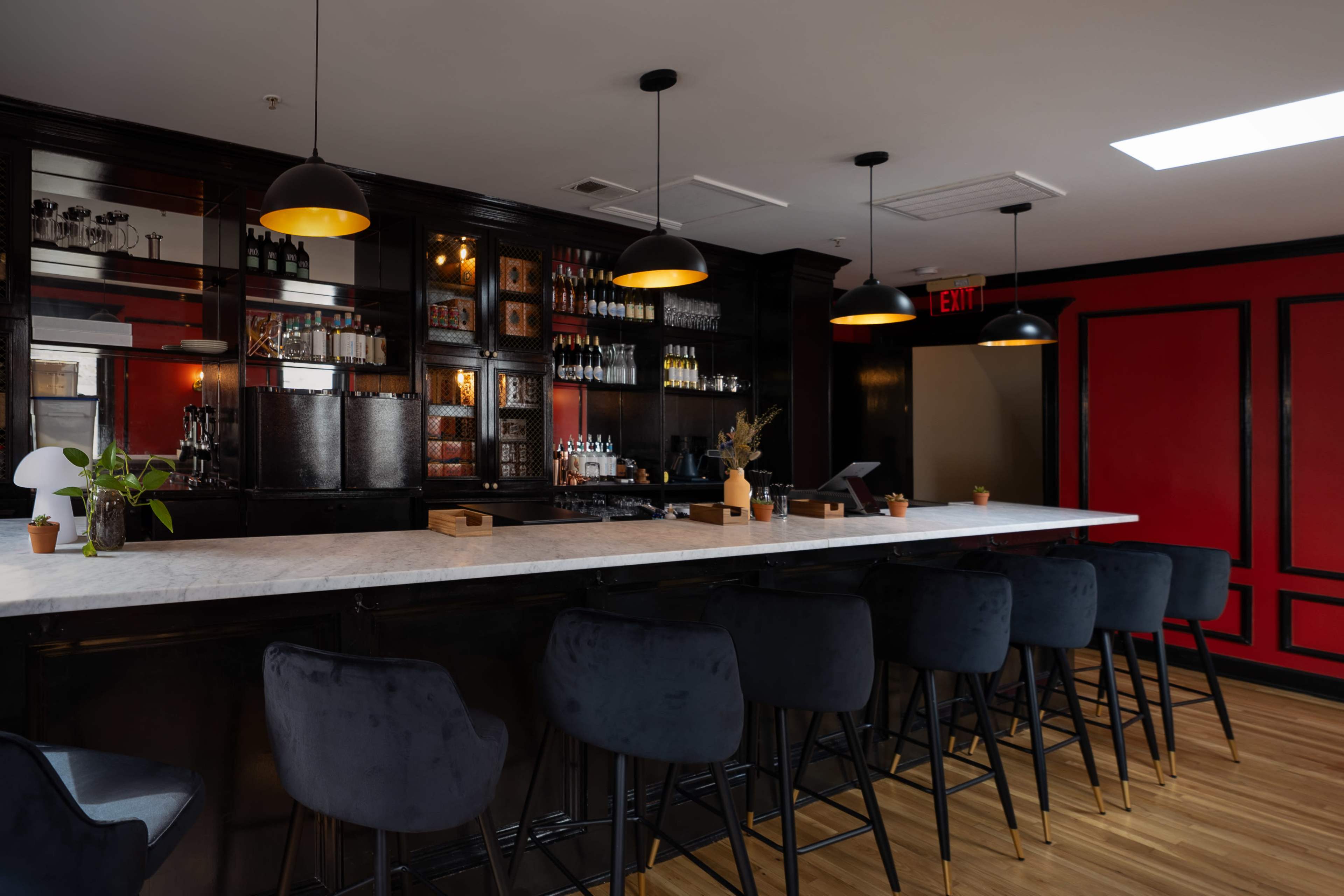A modern bar area features a marble countertop with black upholstered stools, shelves stocked with beverages, and warm pendant lighting against a red accent wall.