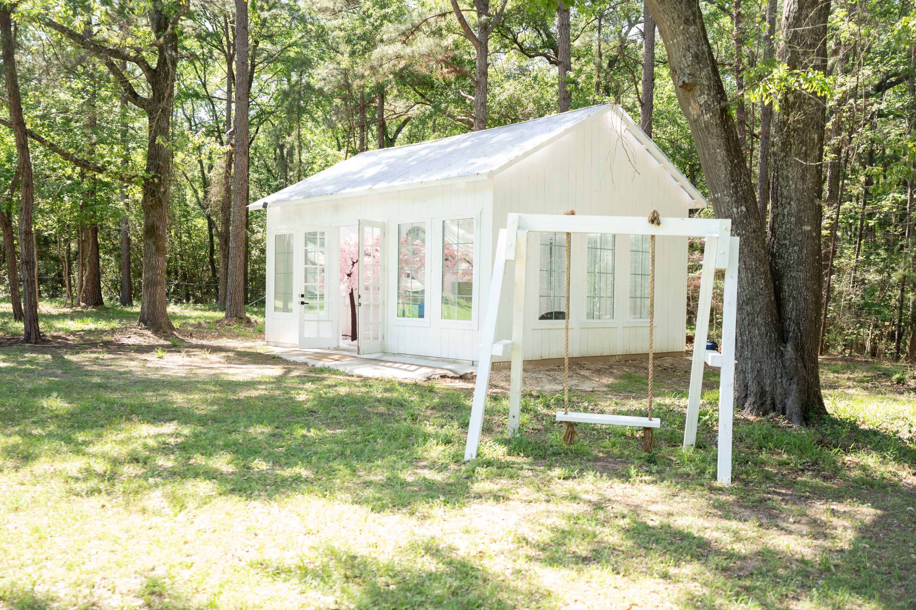 A small white cottage with large windows sits beside a swing in a wooded area.