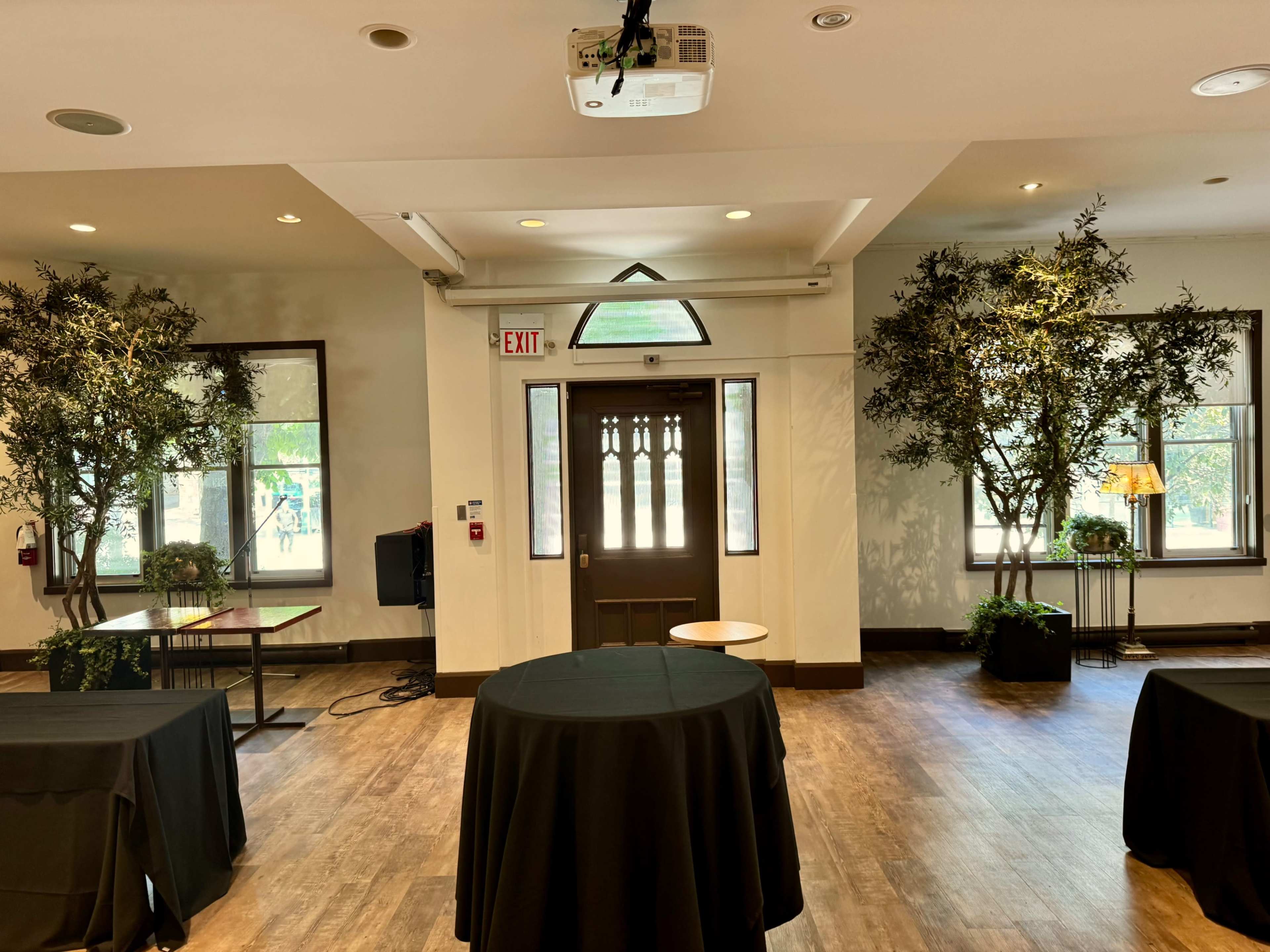 The image shows an indoor event space with two large trees flanking the entrance, black tablecloths on round tables, and a projector mounted on the ceiling.