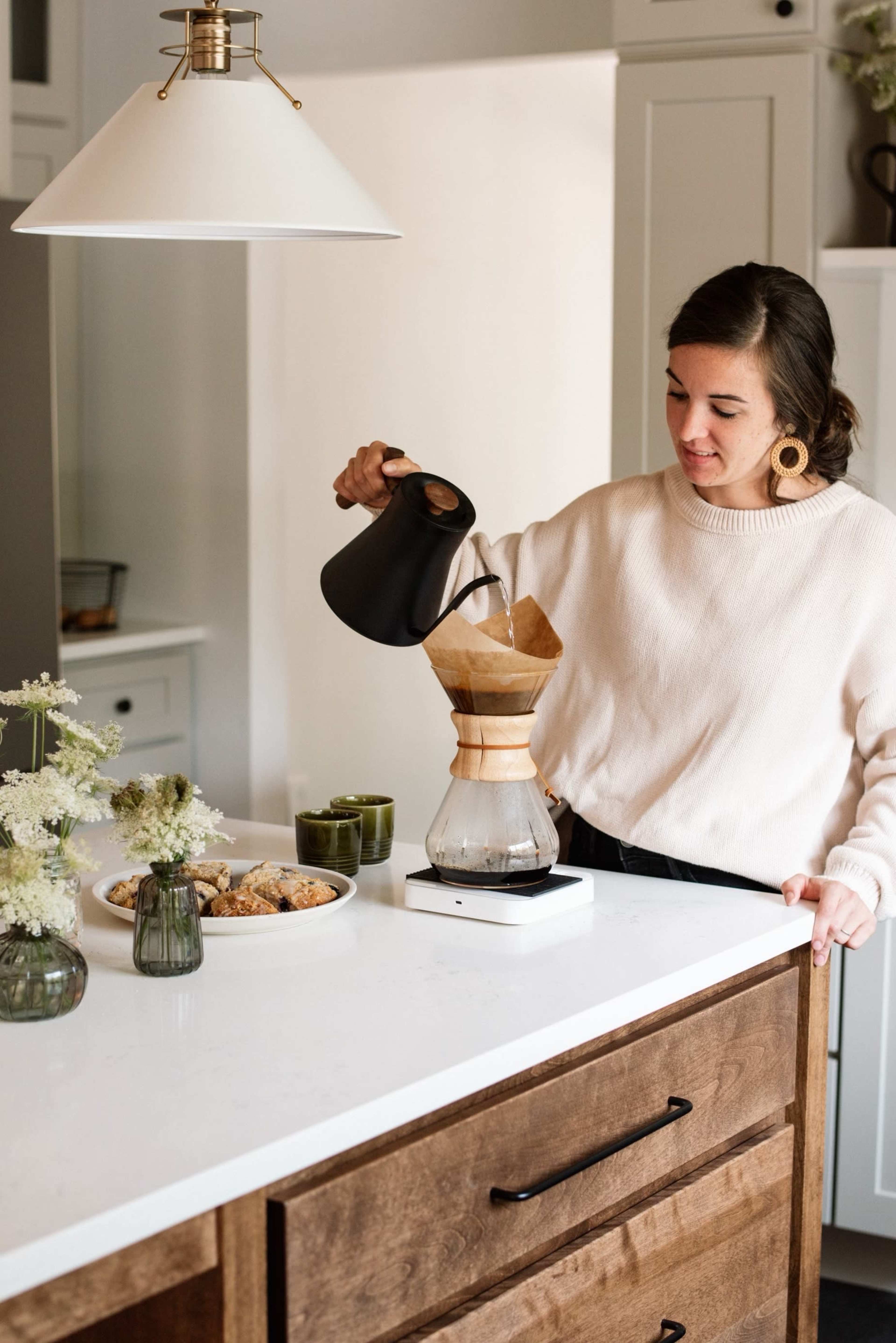 A woman pours hot water from a black kettle into a coffee dripper set on a countertop, with cookies and flowers nearby.