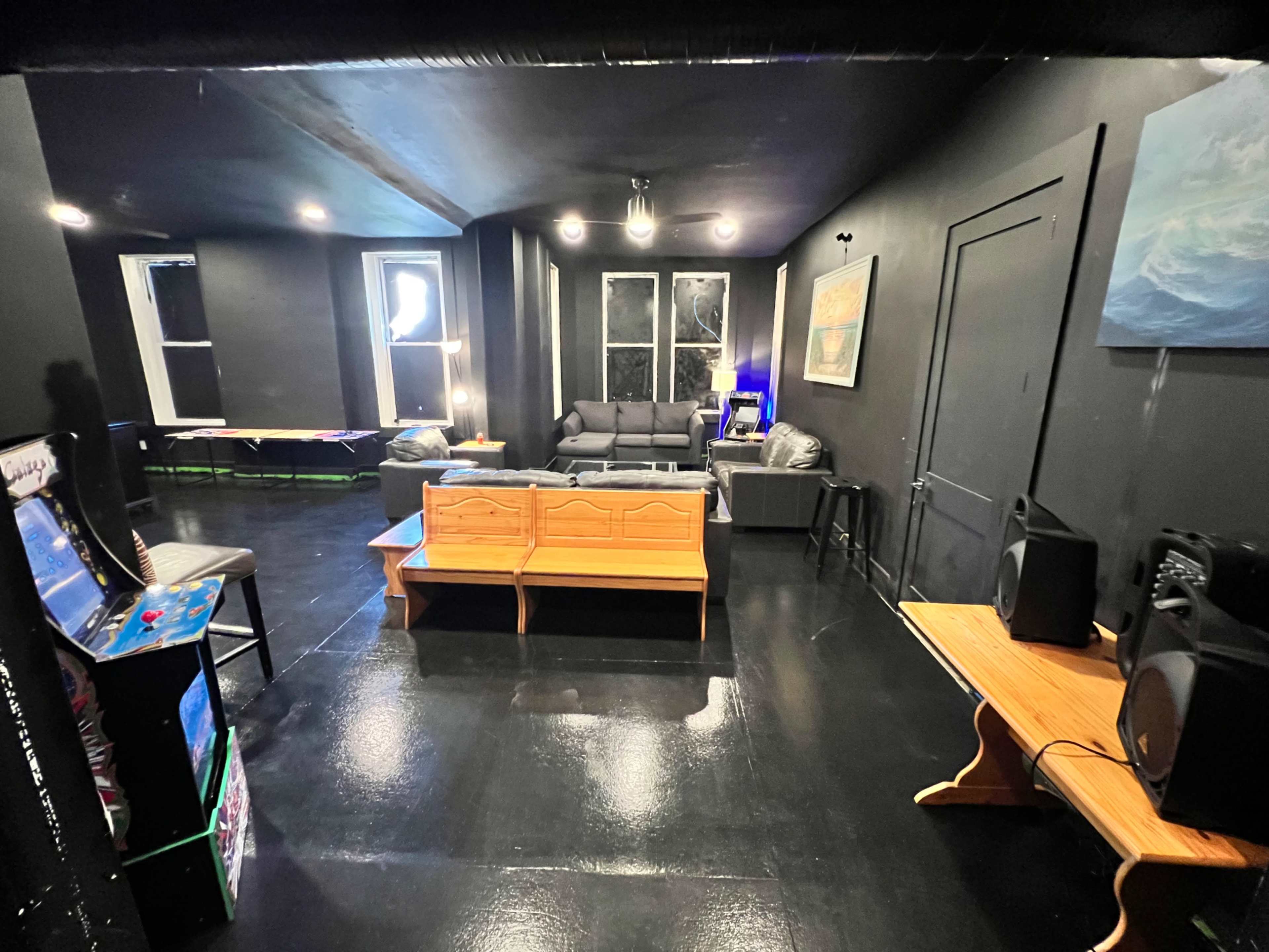 A dimly lit room with black walls, featuring a sofa, two wooden coffee tables, and speakers, alongside an arcade machine in one corner.
