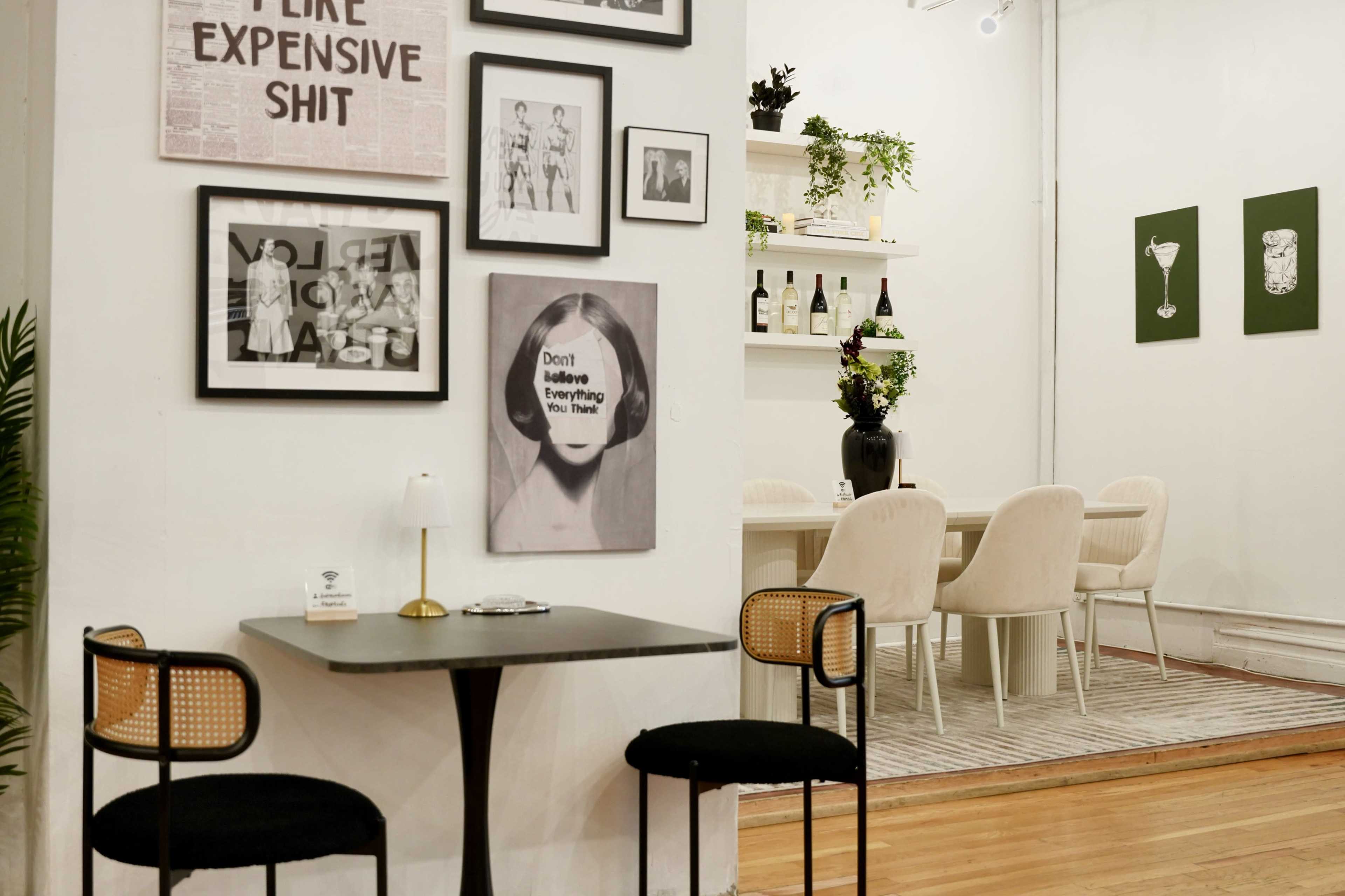 The image shows a cozy café interior featuring a small black table with two chairs, framed artwork on the walls, and a dining area with a large table surrounded by beige chairs.