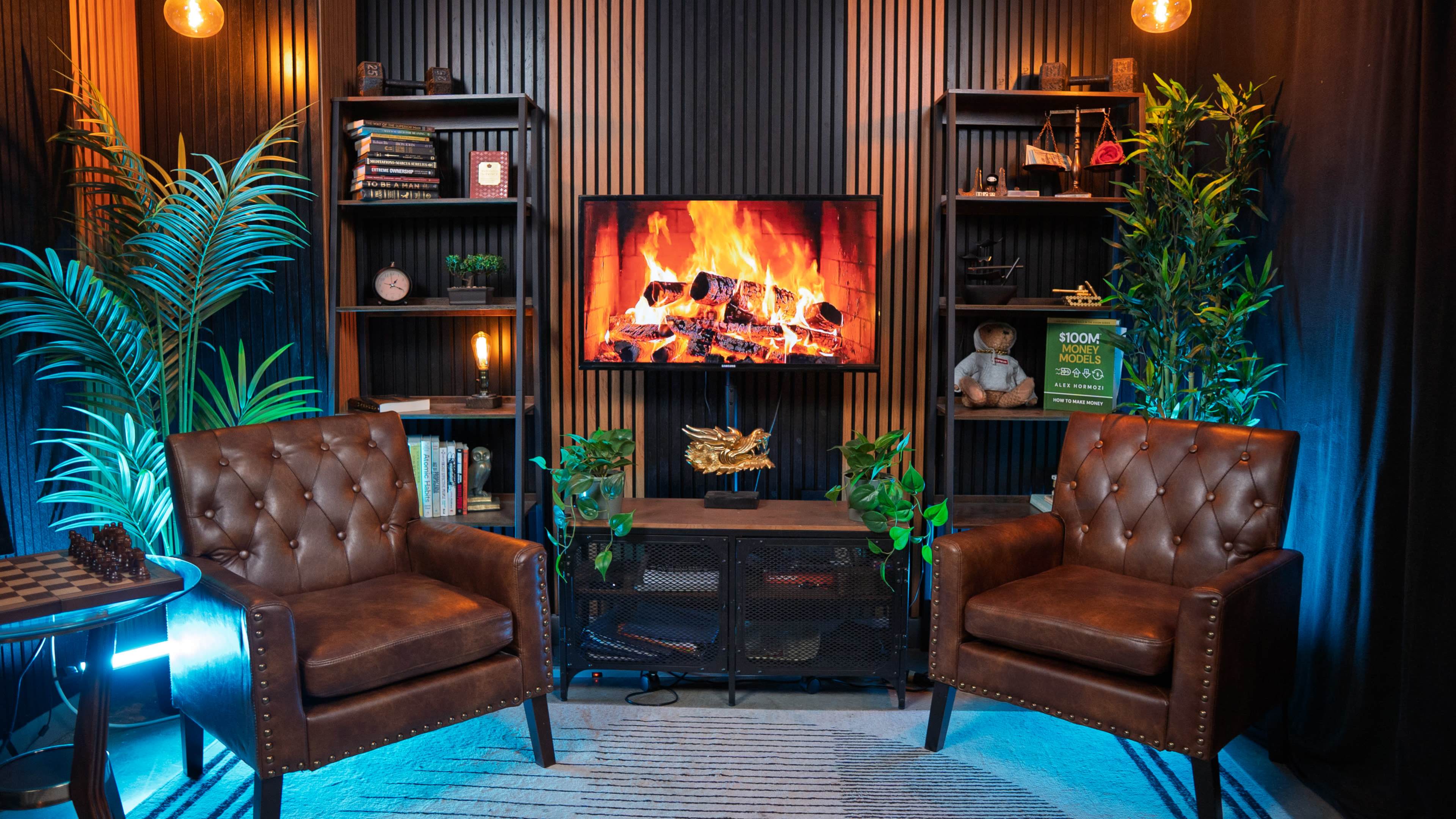 The image depicts a cozy living room setup featuring two brown leather armchairs facing a television displaying a virtual fireplace, flanked by green plants and bookshelves.