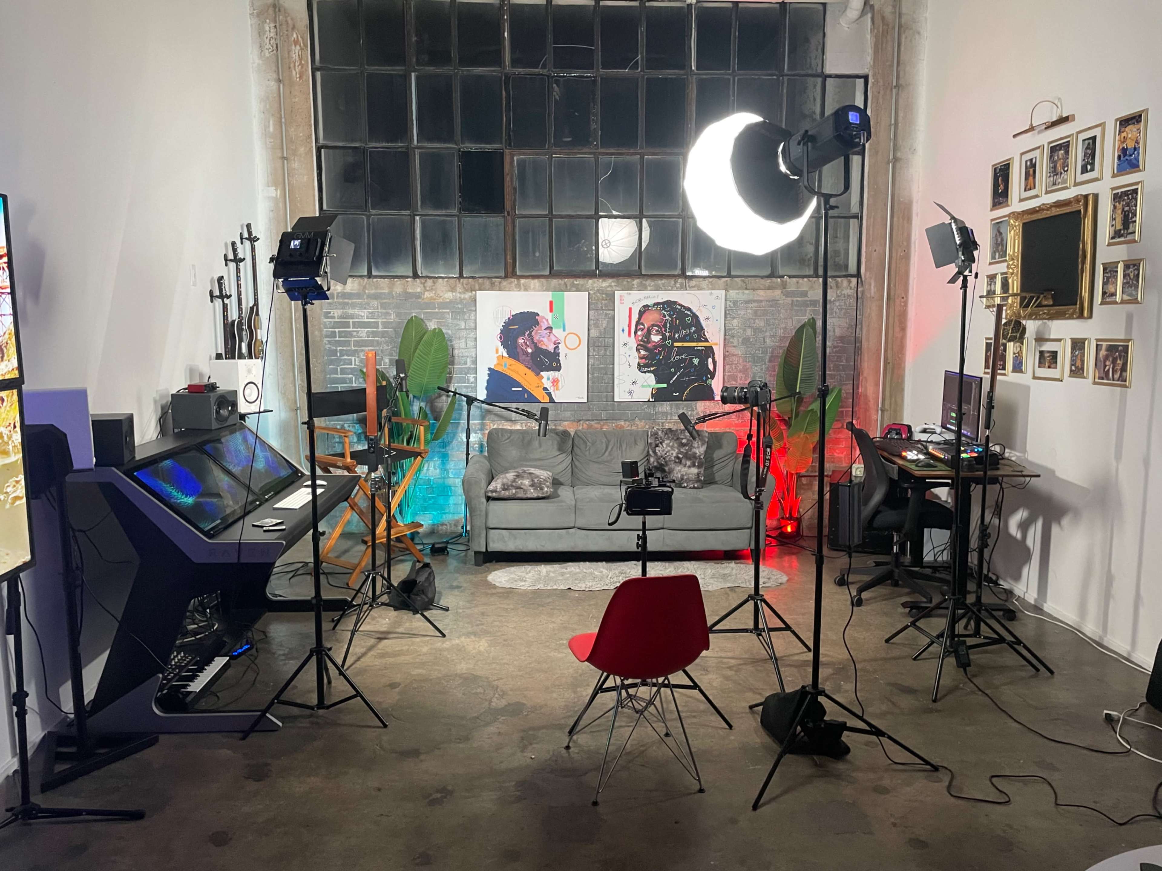 A well-lit studio space with two art pieces on the wall, a gray couch in the center, and equipment set up for video production, including cameras and lights.