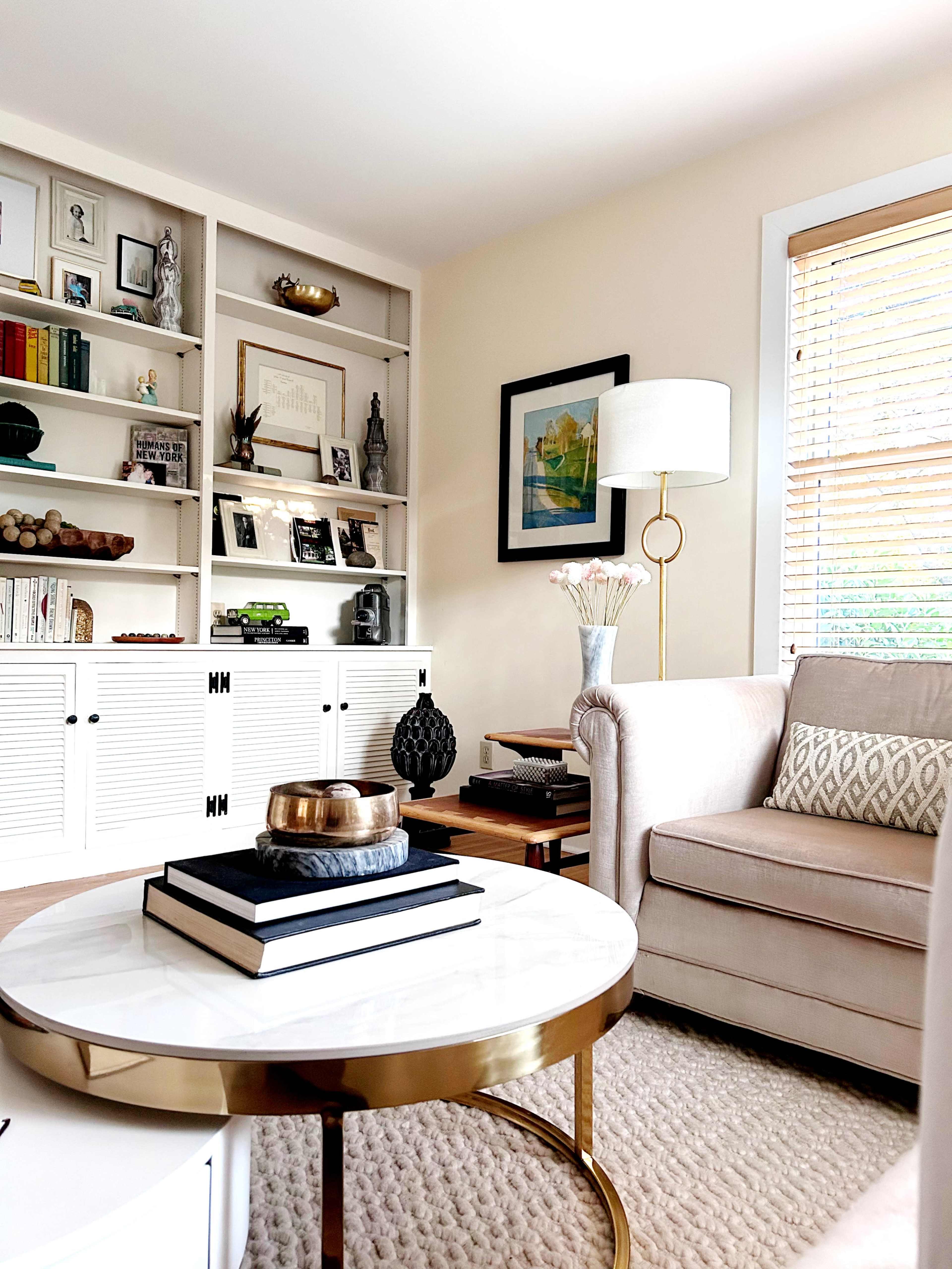The image depicts a stylish living room featuring a beige sofa, a circular coffee table with a marble top and gold accents, and bookshelves filled with various decorative items.