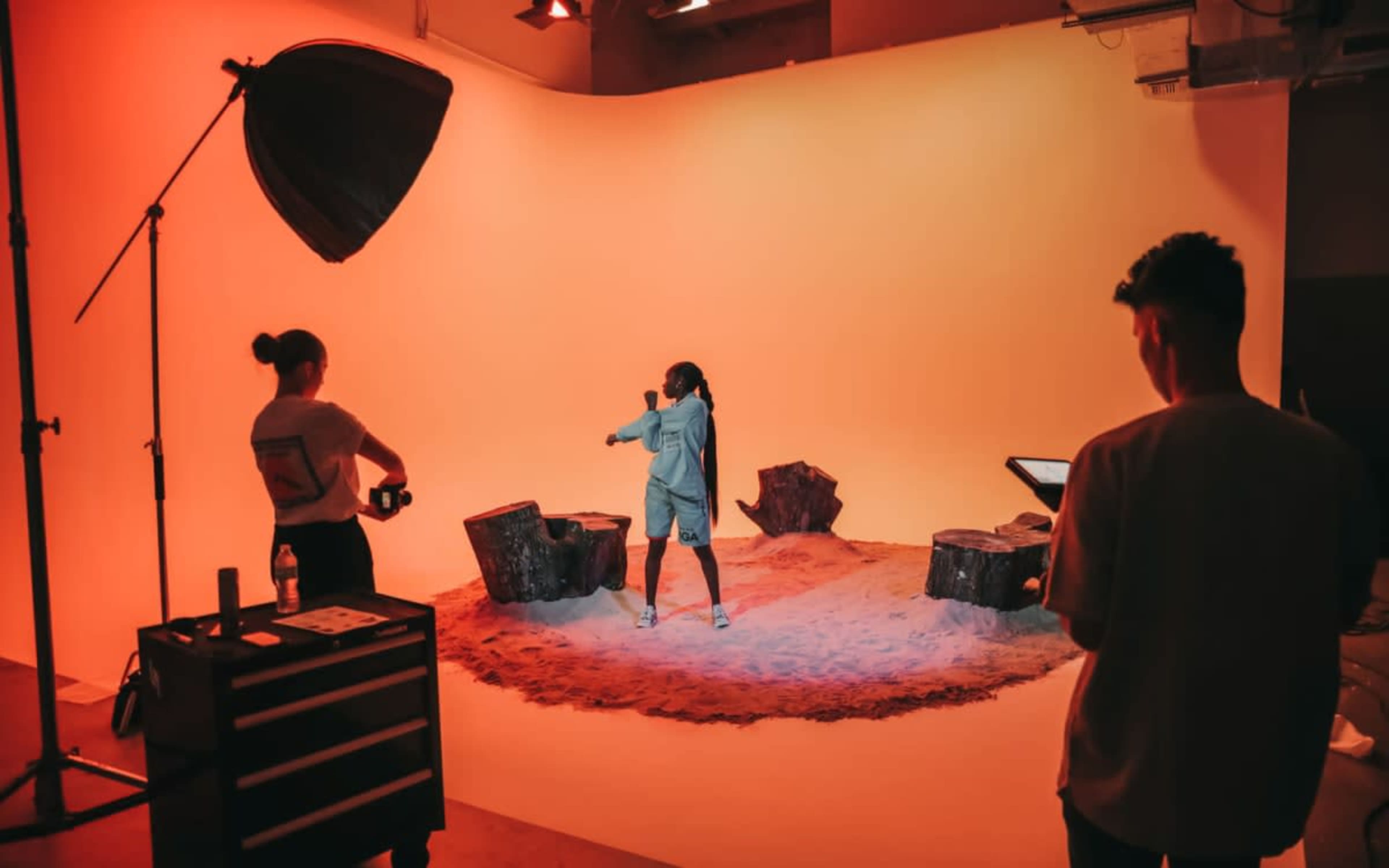 A model poses in front of an orange backdrop in a photography studio, while a crew member sets up equipment nearby.